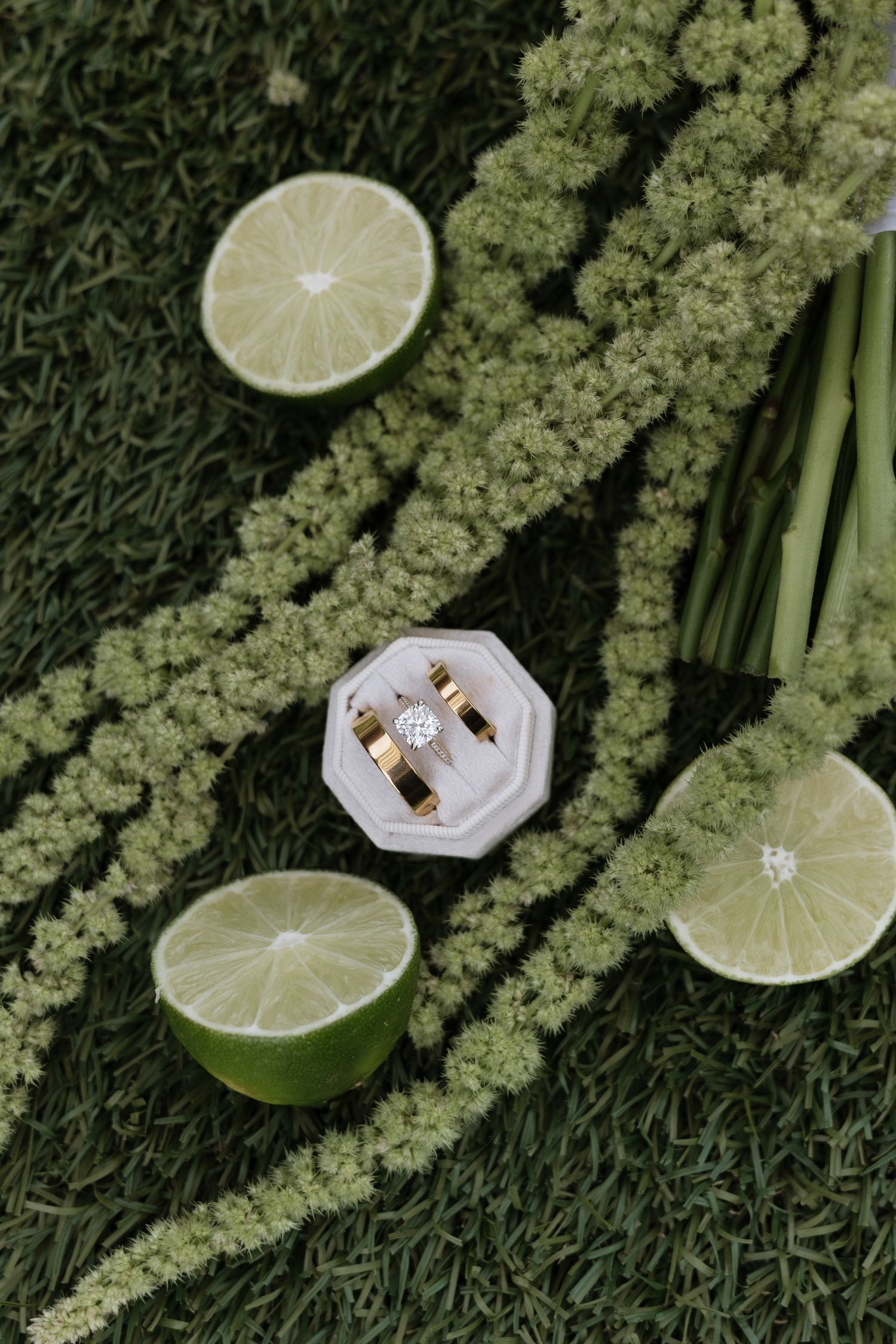 Gold wedding rings in a white octagonal box surrounded by halved limes and green floral stems on grass.