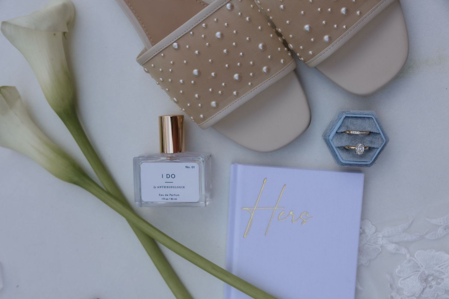 Bridal flat lay with calla lilies, perfume, rings in blue velvet box, sandals, and vow book.