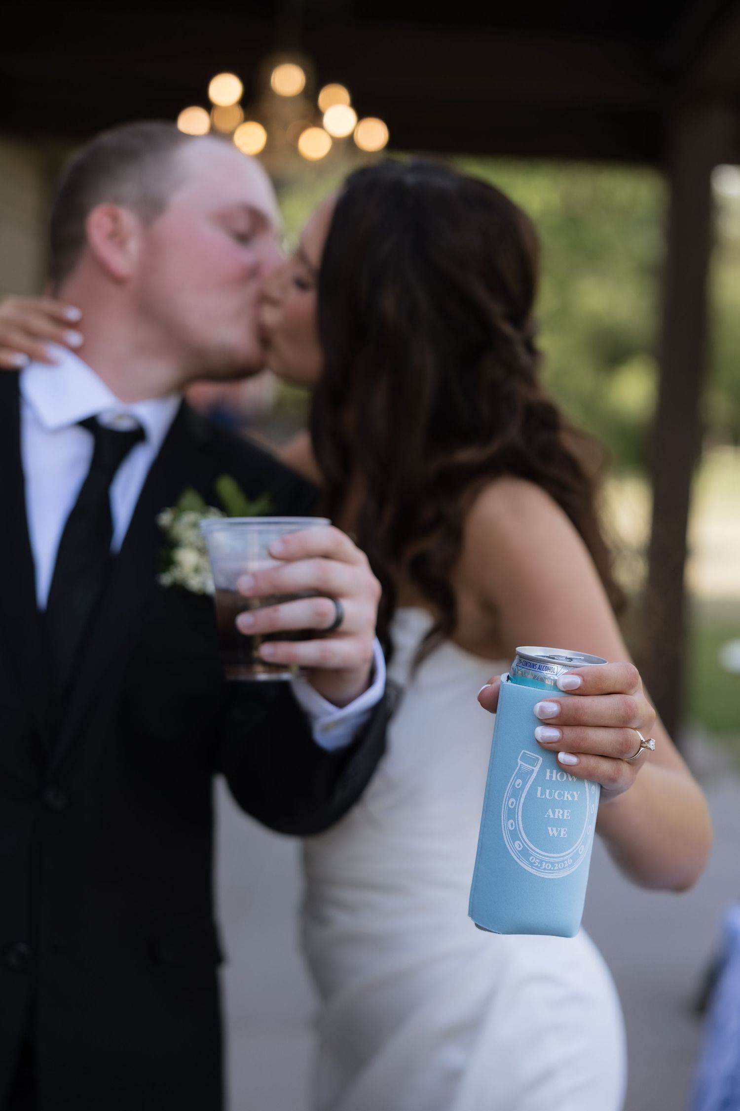 Wedding couple kisses while bride holds a custom light blue can koozie at outdoor reception.