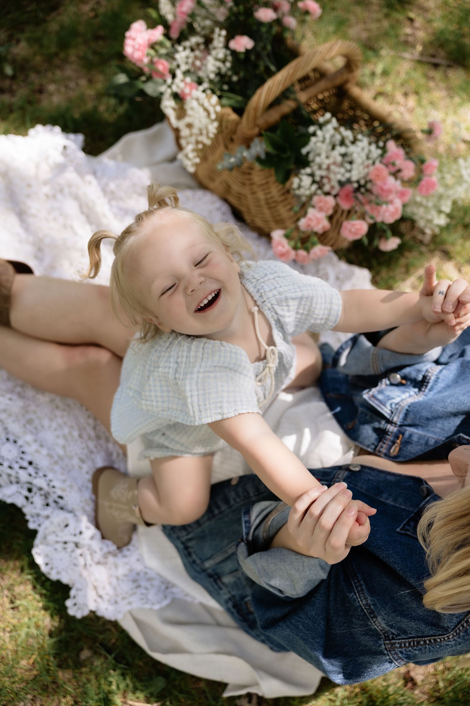 Laughing toddler girl lies on picnic blanket outdoors surrounded by flowers and family members.
