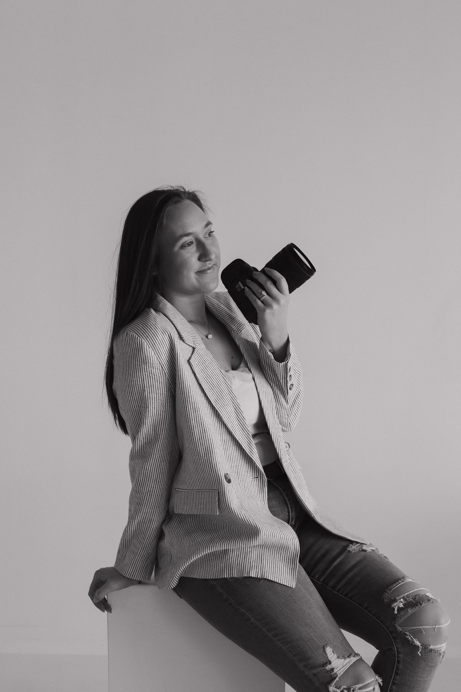 Black and white portrait of smiling female photographer sitting on cube holding camera in blazer.