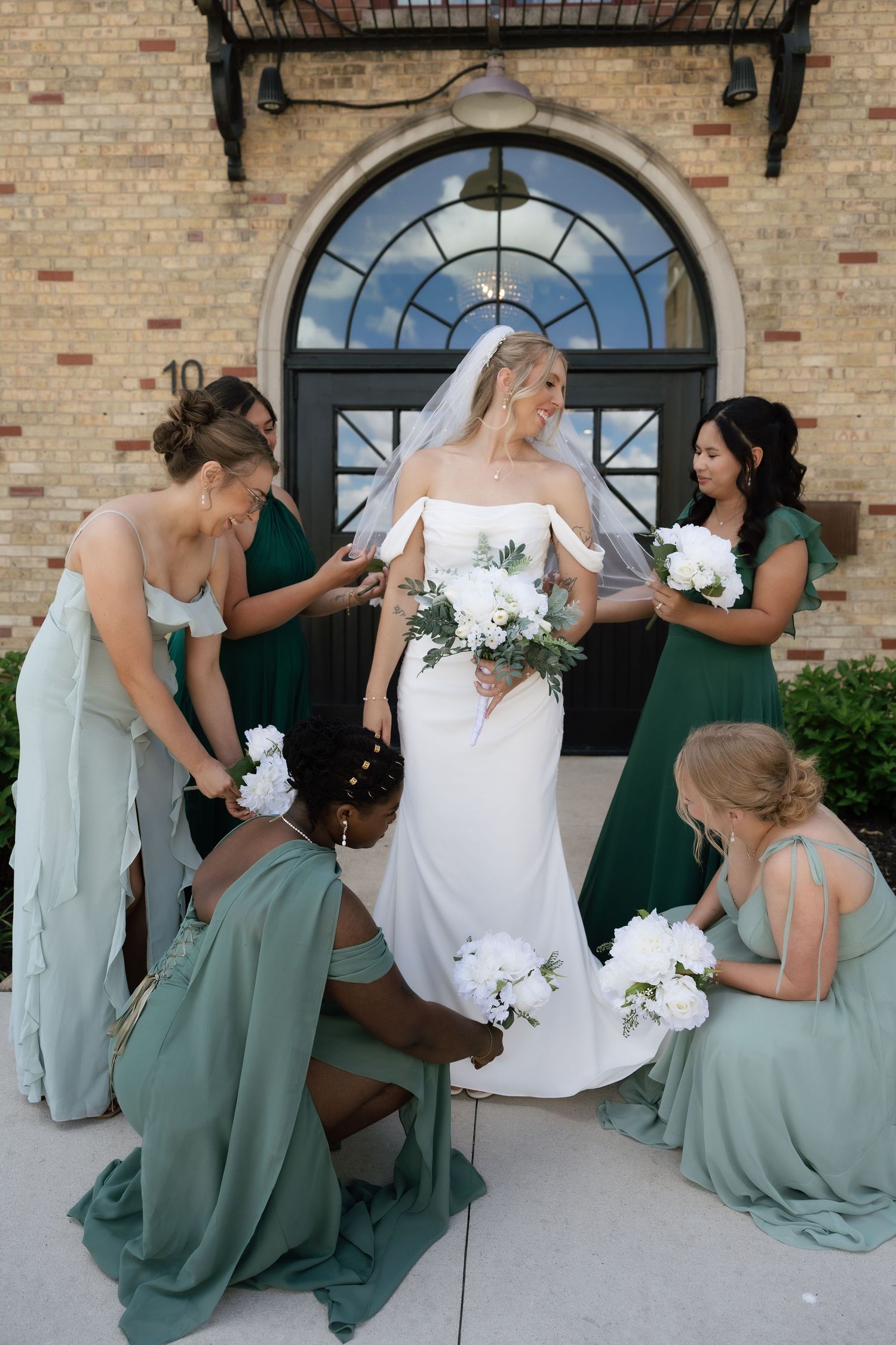 Bride in white gown holds bouquet while bridesmaids in sage green dresses assist her outside brick building.