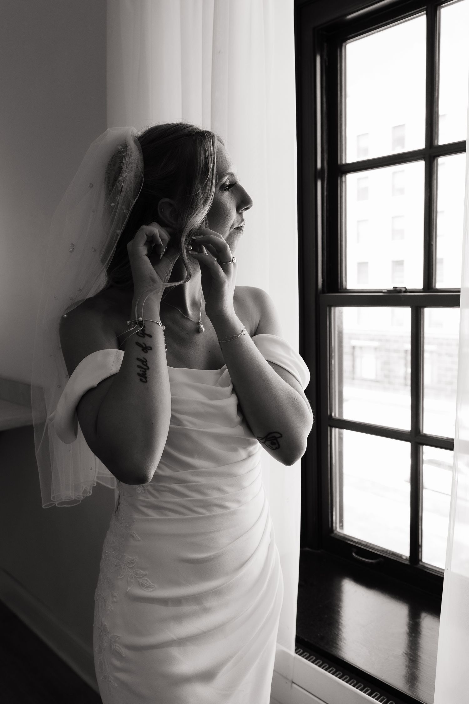Black and white photo of a bride in a white gown and veil adjusting her earring by a window.