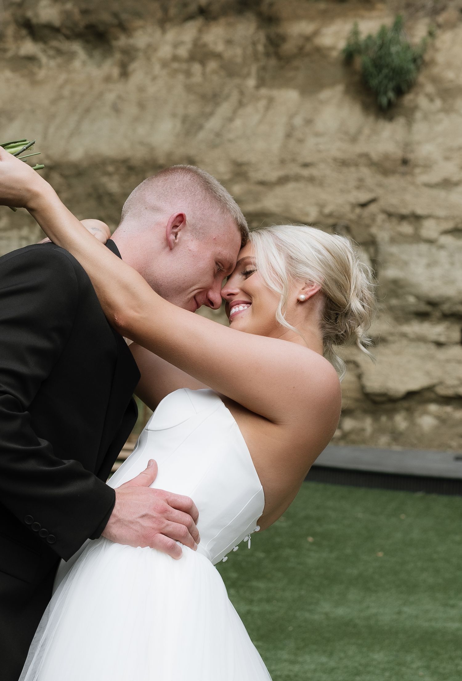 Bride and groom sharing a romantic embrace, smiling together against a rustic stone wall backdrop.