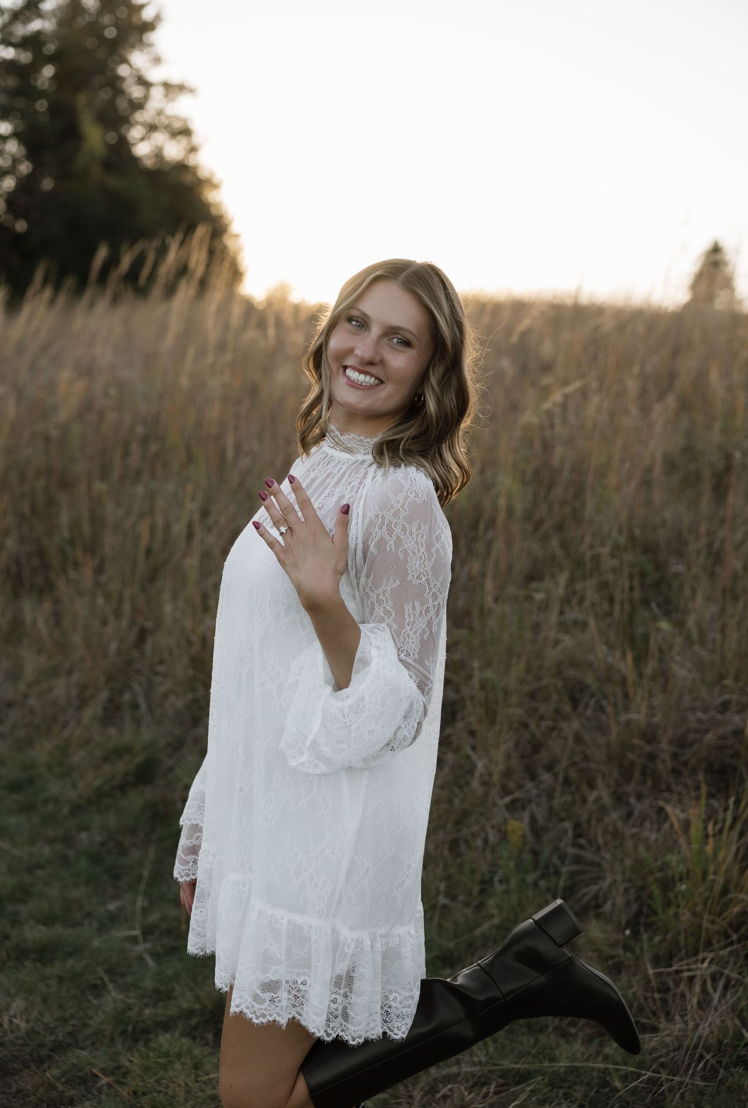 Young woman in white lace dress and black boots smiling in a golden field at sunset.