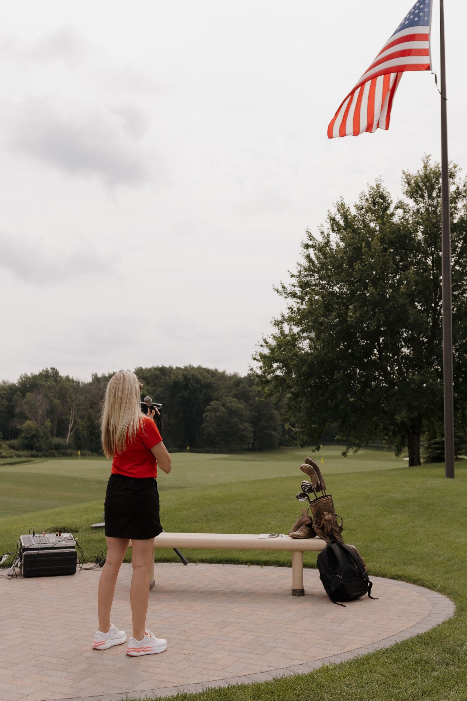 Blonde woman in red top photographing a golf bag on a bench near an American flag on a lush green golf course.