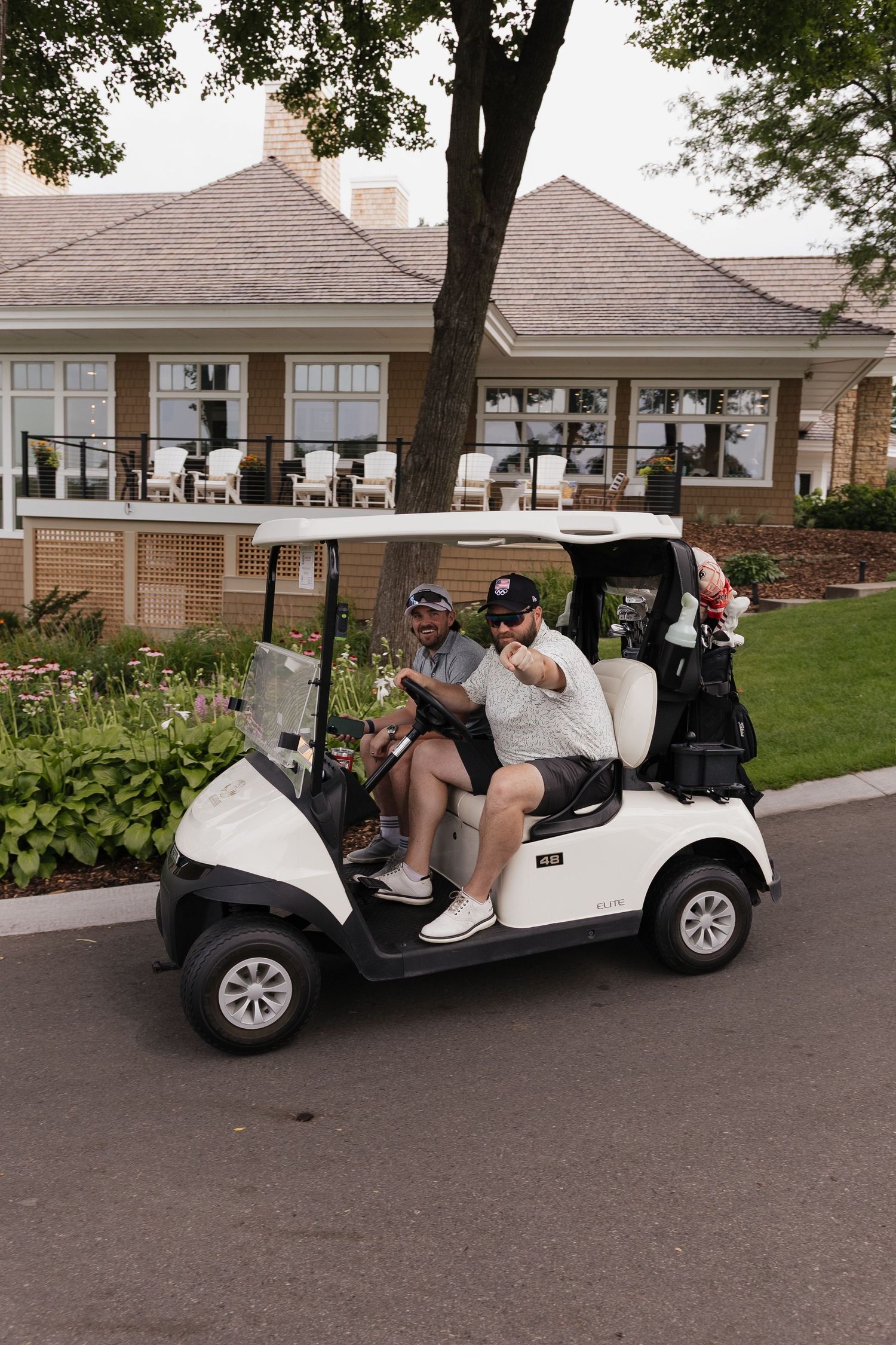 Two men riding a white golf cart near a clubhouse, one giving a thumbs up on a sunny day.