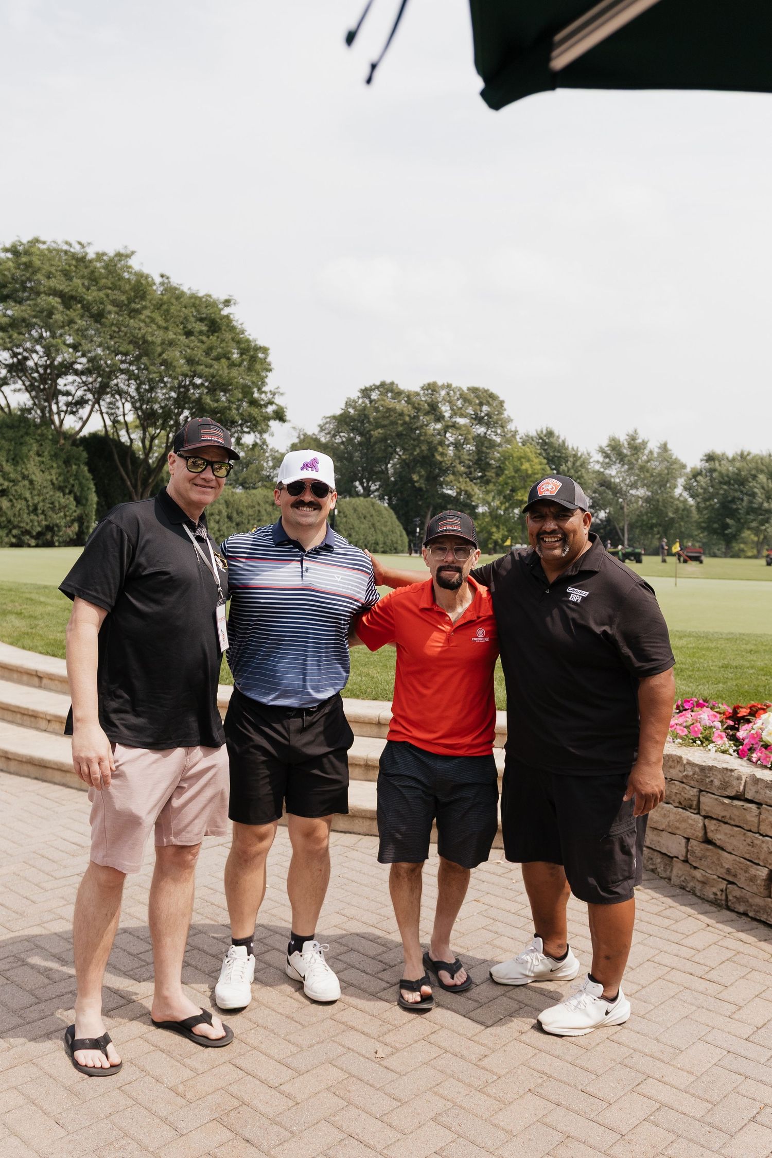 Four men posing together on a golf course patio on a sunny day, wearing casual golf attire and caps.