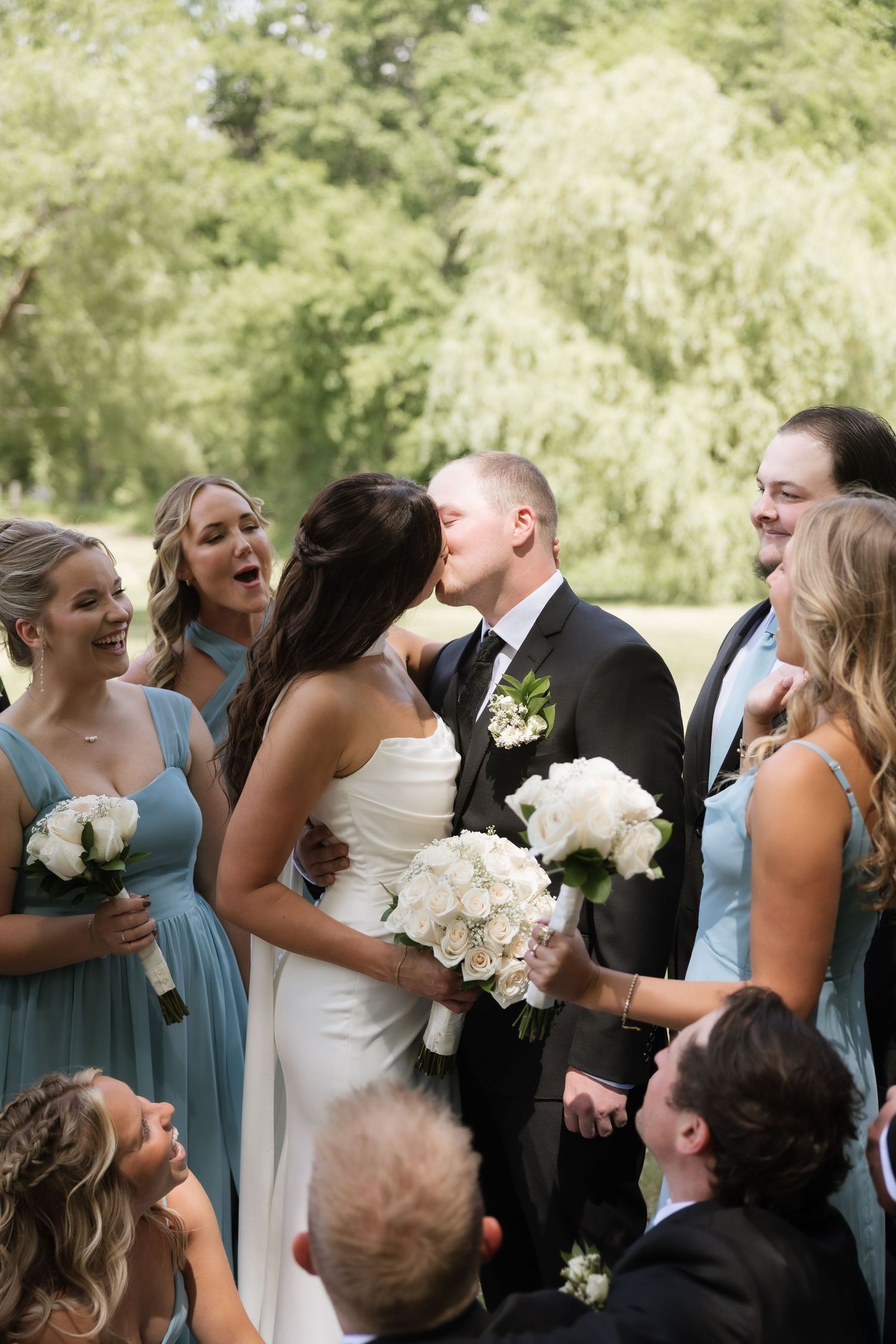 Bride and groom share a kiss surrounded by a cheering wedding party in dusty blue bridesmaid dresses outdoors.