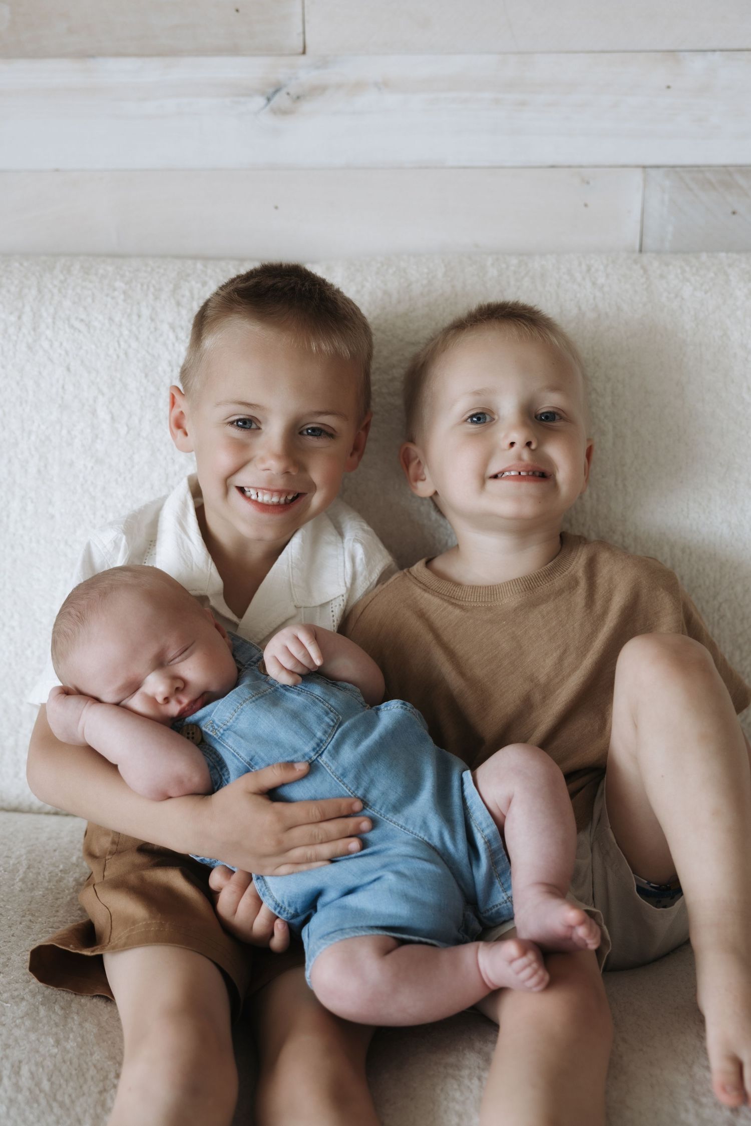 Two smiling young brothers sitting together holding their sleeping newborn sibling in blue romper.
