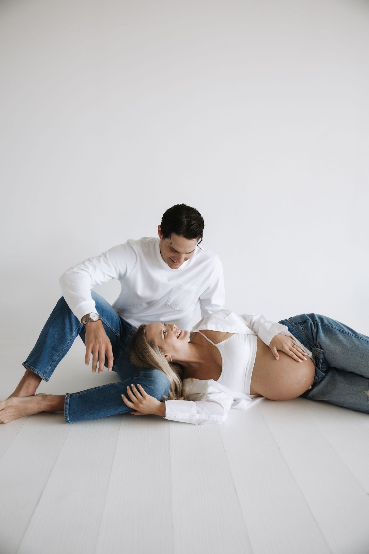 Couple in matching white tops and jeans relaxing on white floor, man gazing at pregnant partner lovingly.