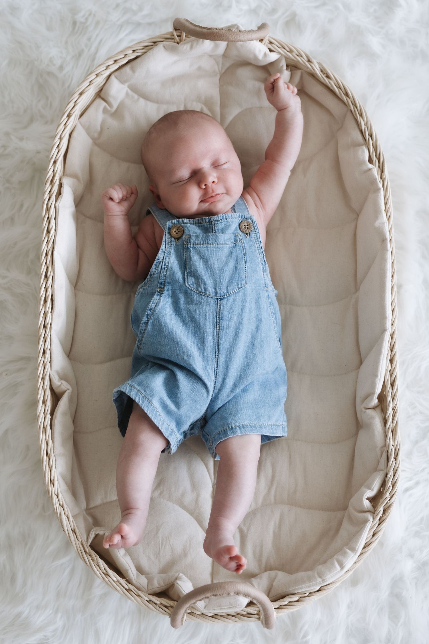Newborn baby stretching arms in wicker Moses basket wearing blue denim overalls on white fur background.