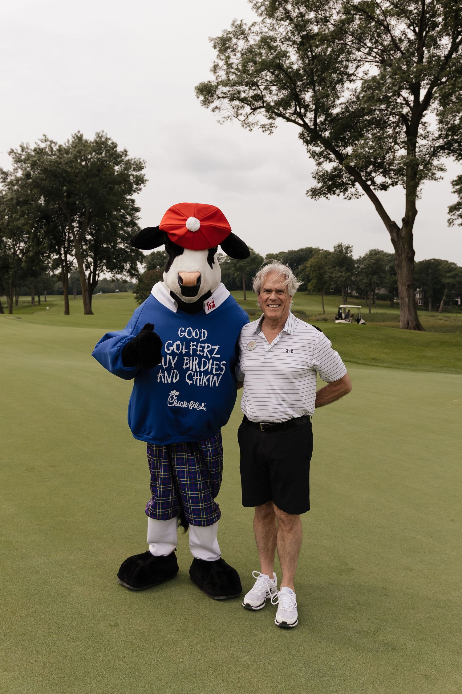 Man poses with Chick-fil-A cow mascot on golf course green, mascot wearing blue sweater with golf-themed message.