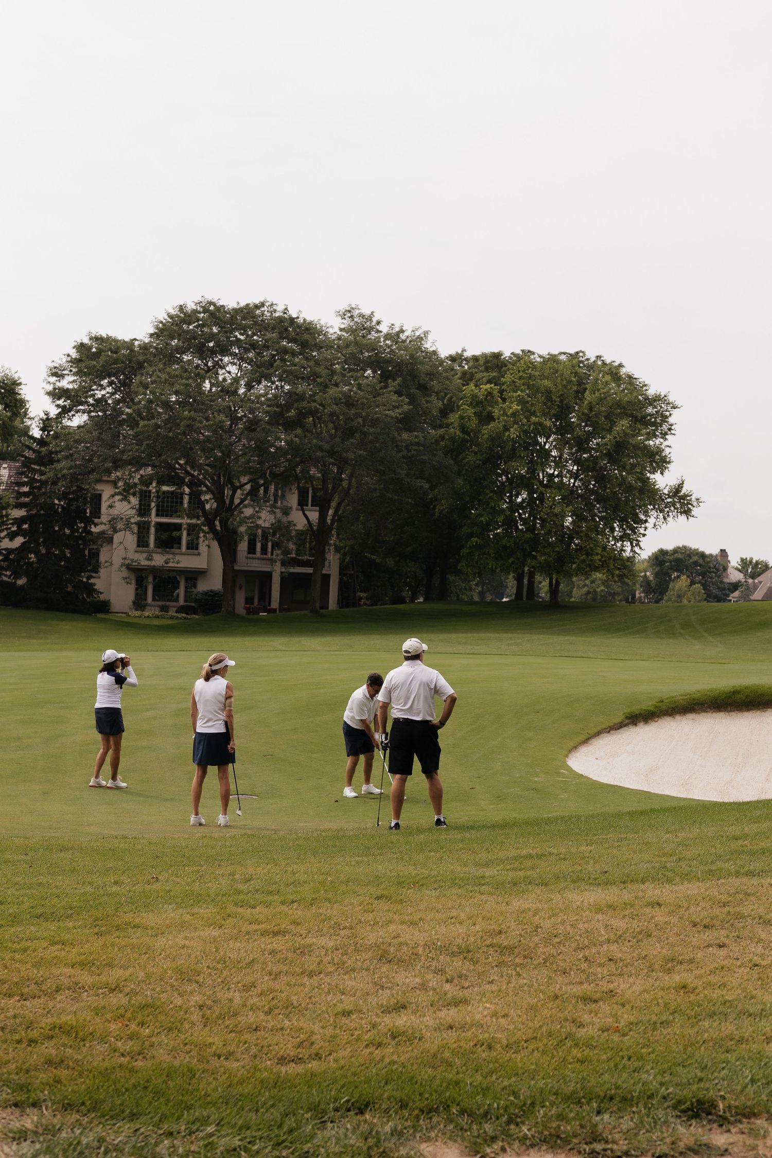 Four golfers on a lush green golf course near a sand bunker, with trees and a clubhouse in the background.