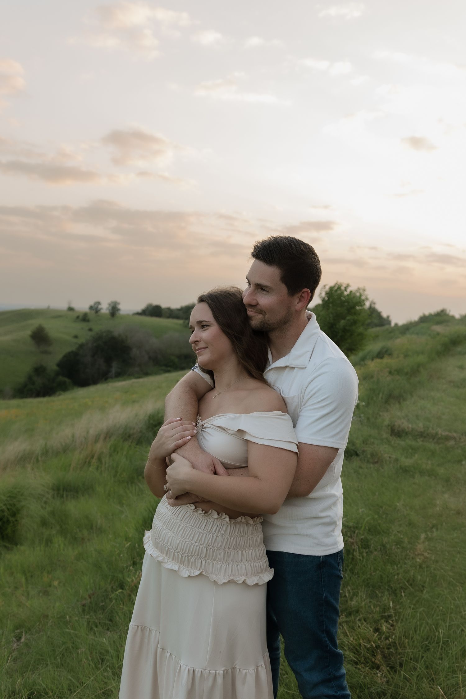 A couple embraces at sunset on a green hillside, the woman in a white dress and man in a white polo shirt.