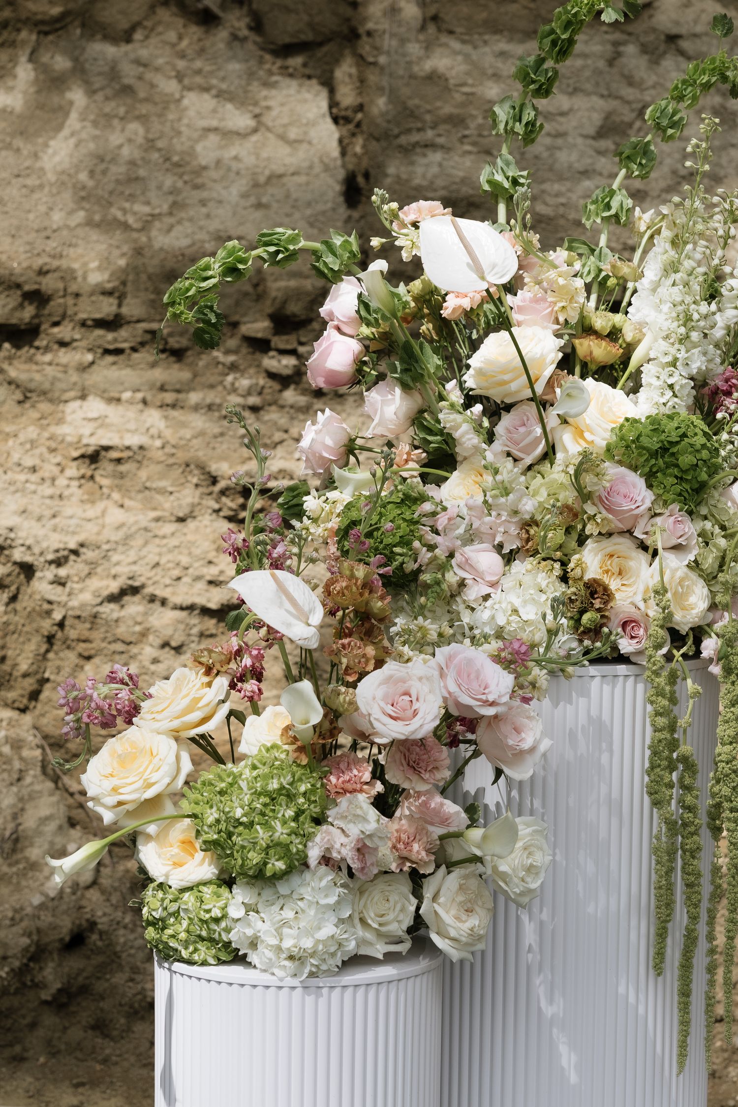 Lush floral arrangement of pink and white roses on white pedestal columns against a stone wall.