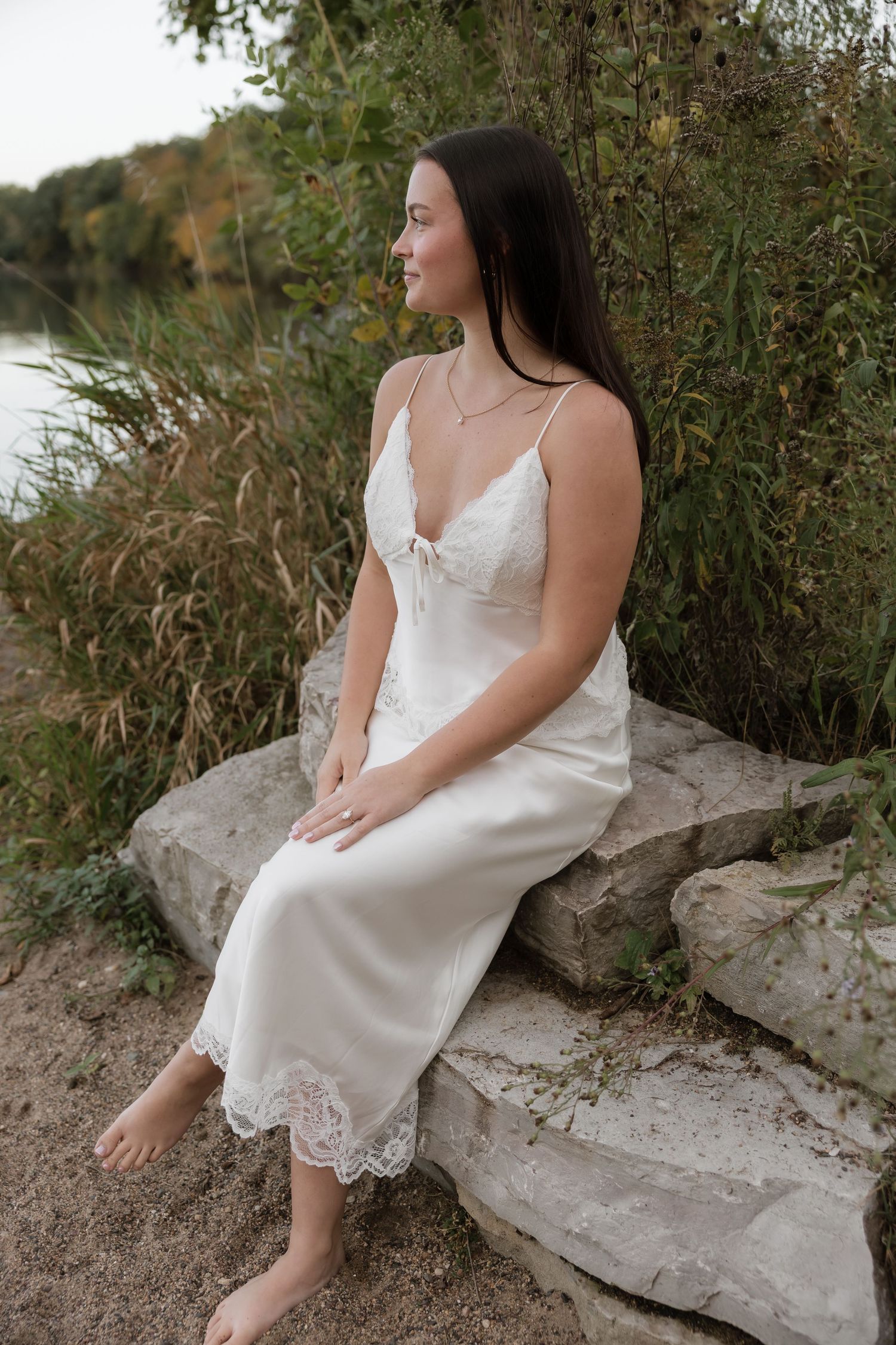 Bride in white lace slip dress sitting barefoot on stone steps by a river surrounded by tall grass.