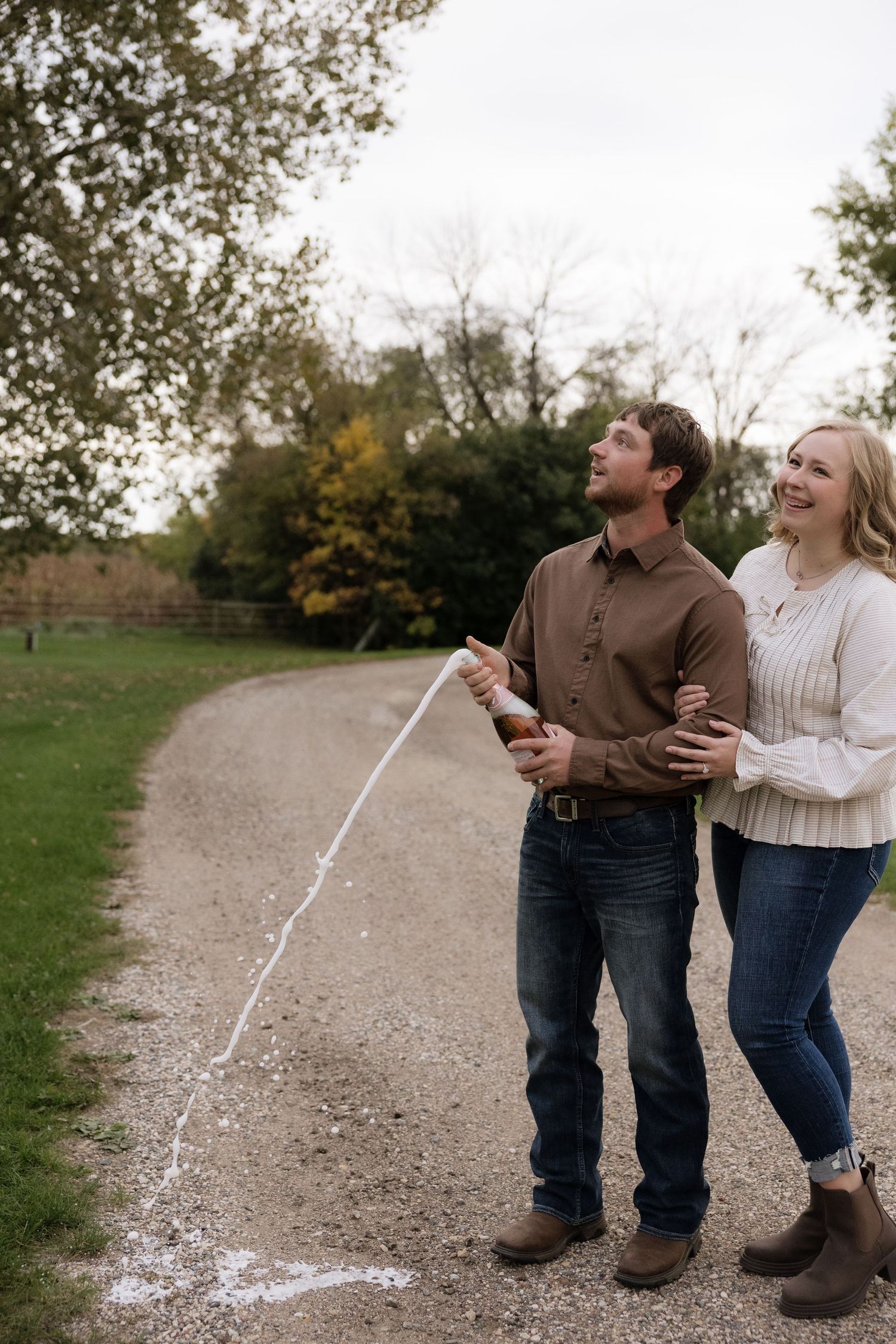 Happy couple popping champagne on a gravel path surrounded by autumn trees during an outdoor celebration.