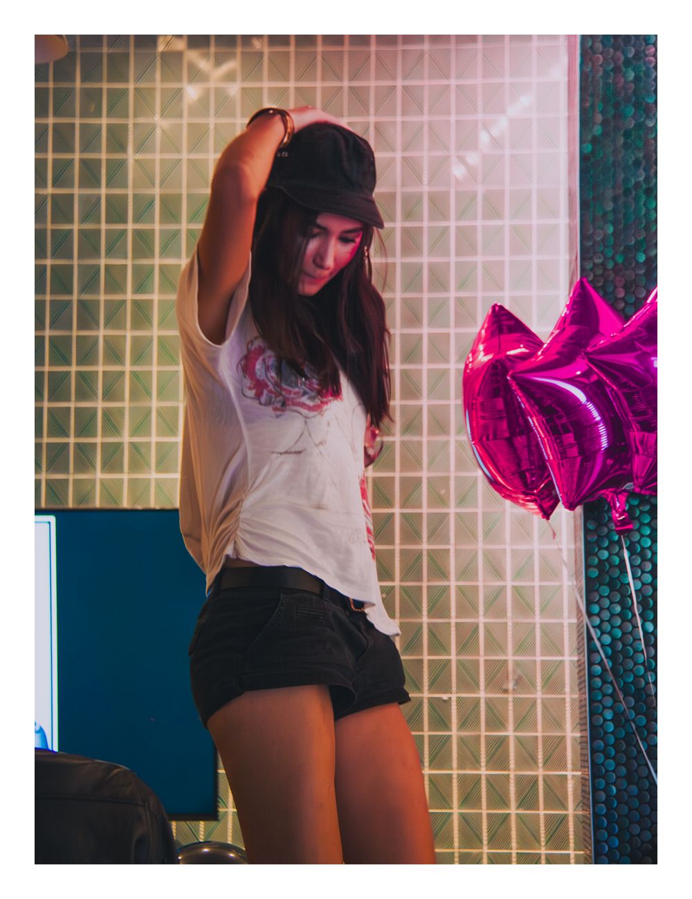 Young woman in white graphic tee and denim shorts posing near pink star-shaped balloons indoors.