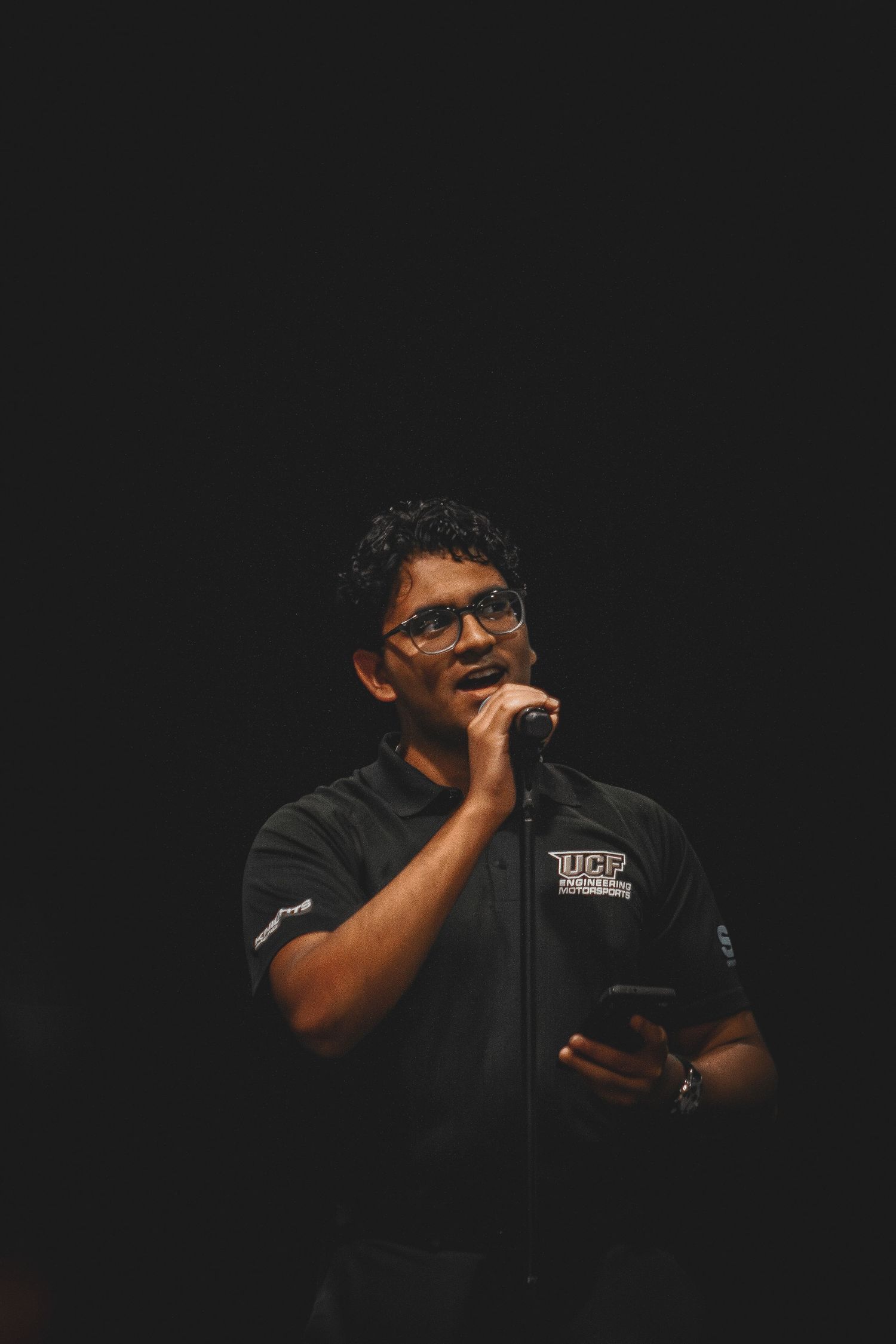 A man in a UCF shirt speaks into a microphone while holding a phone on a dark stage.