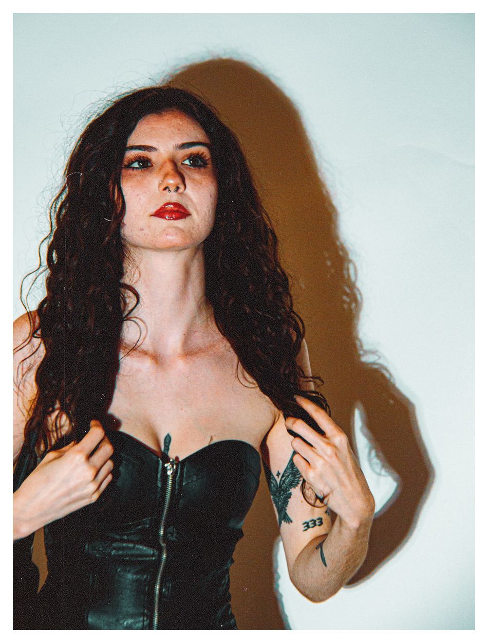 Dark-haired woman in black leather corset with red lips and a rose tattoo poses.