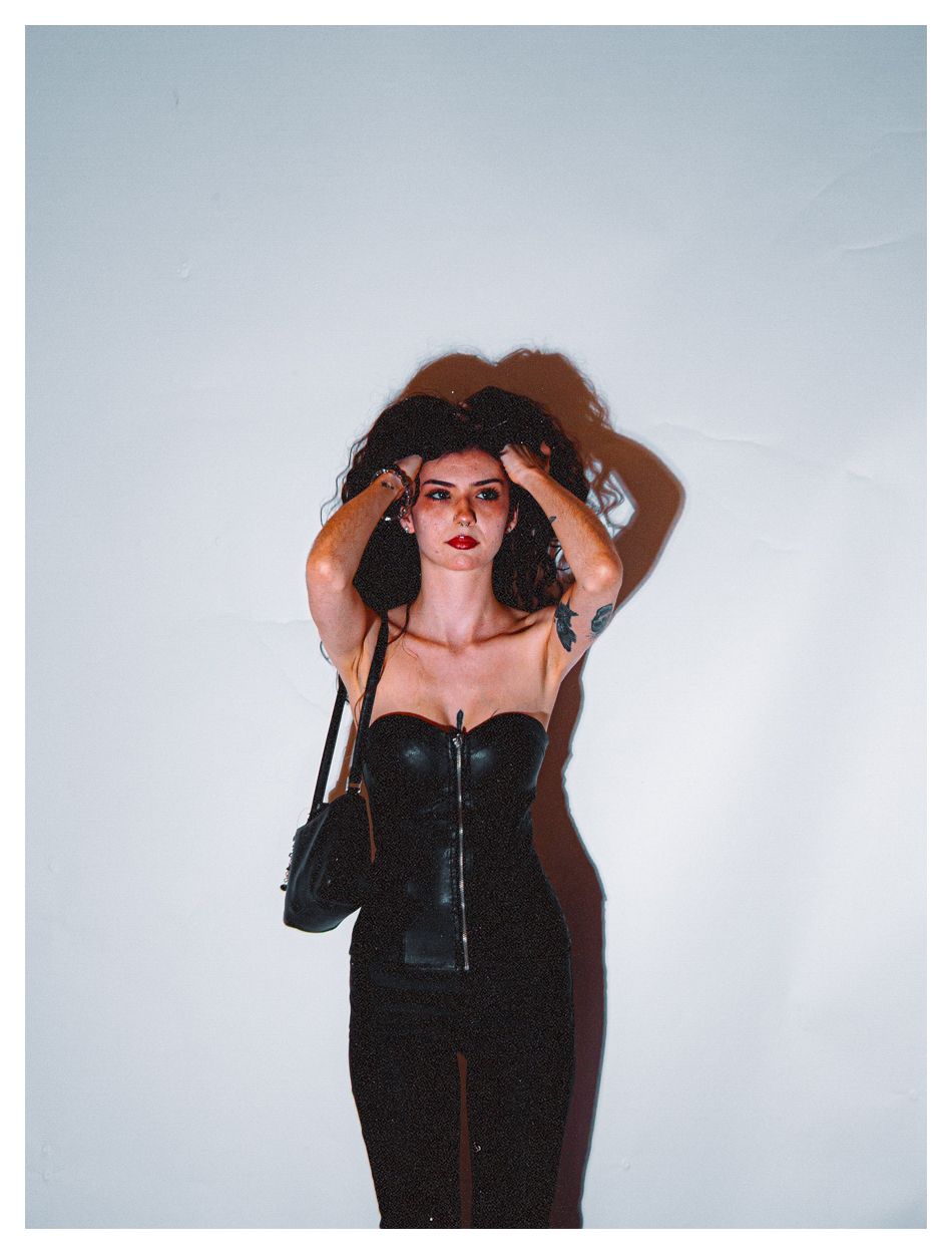Woman in black leather corset dress poses against white wall with hands in hair.
