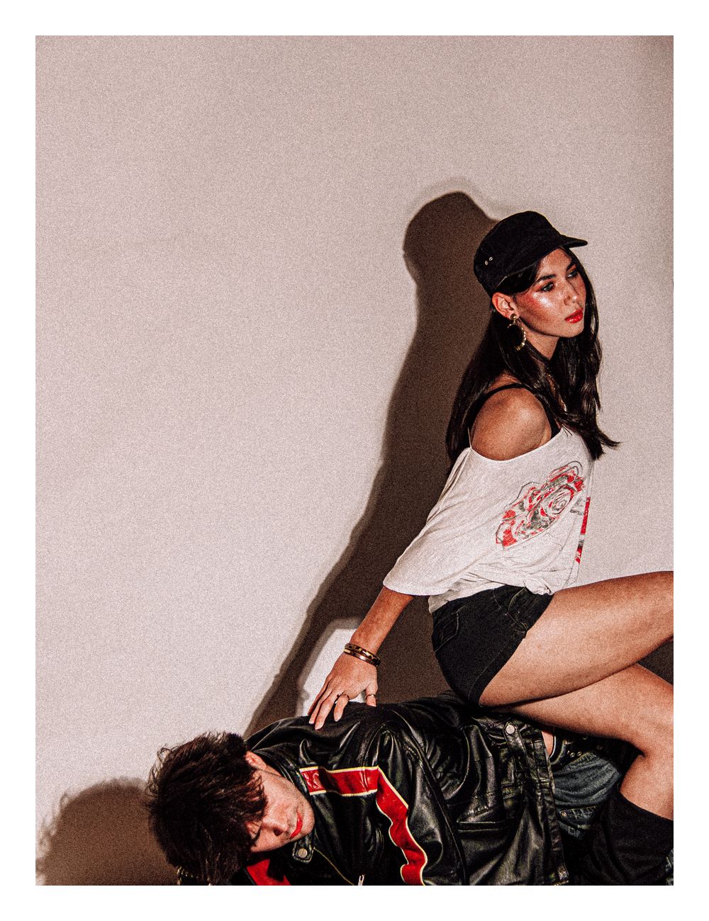 Stylish woman in cap and graphic tee poses over a man in a leather jacket in edgy fashion shoot.
