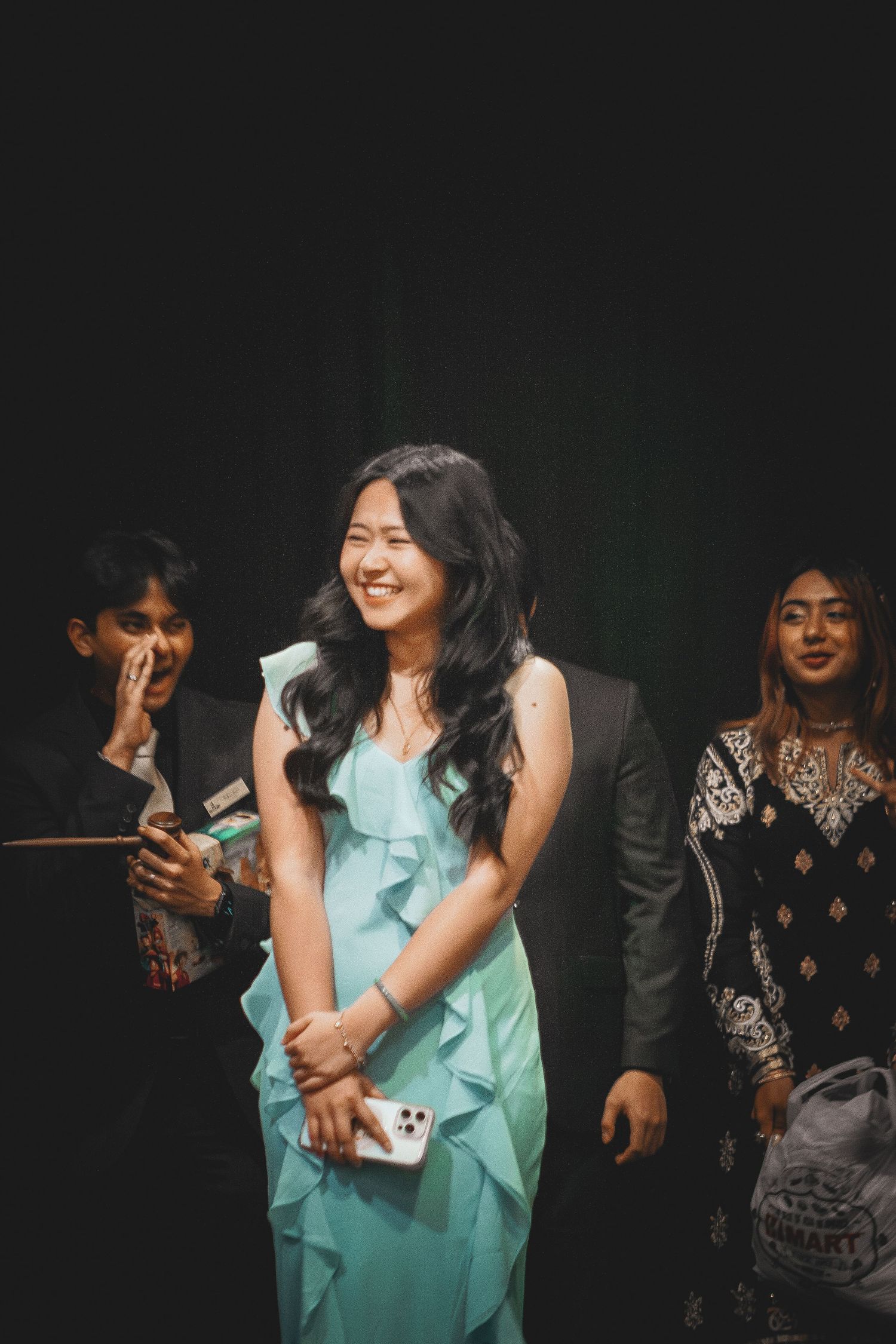 A smiling young woman in a teal ruffled dress stands with others in a dimly lit venue.