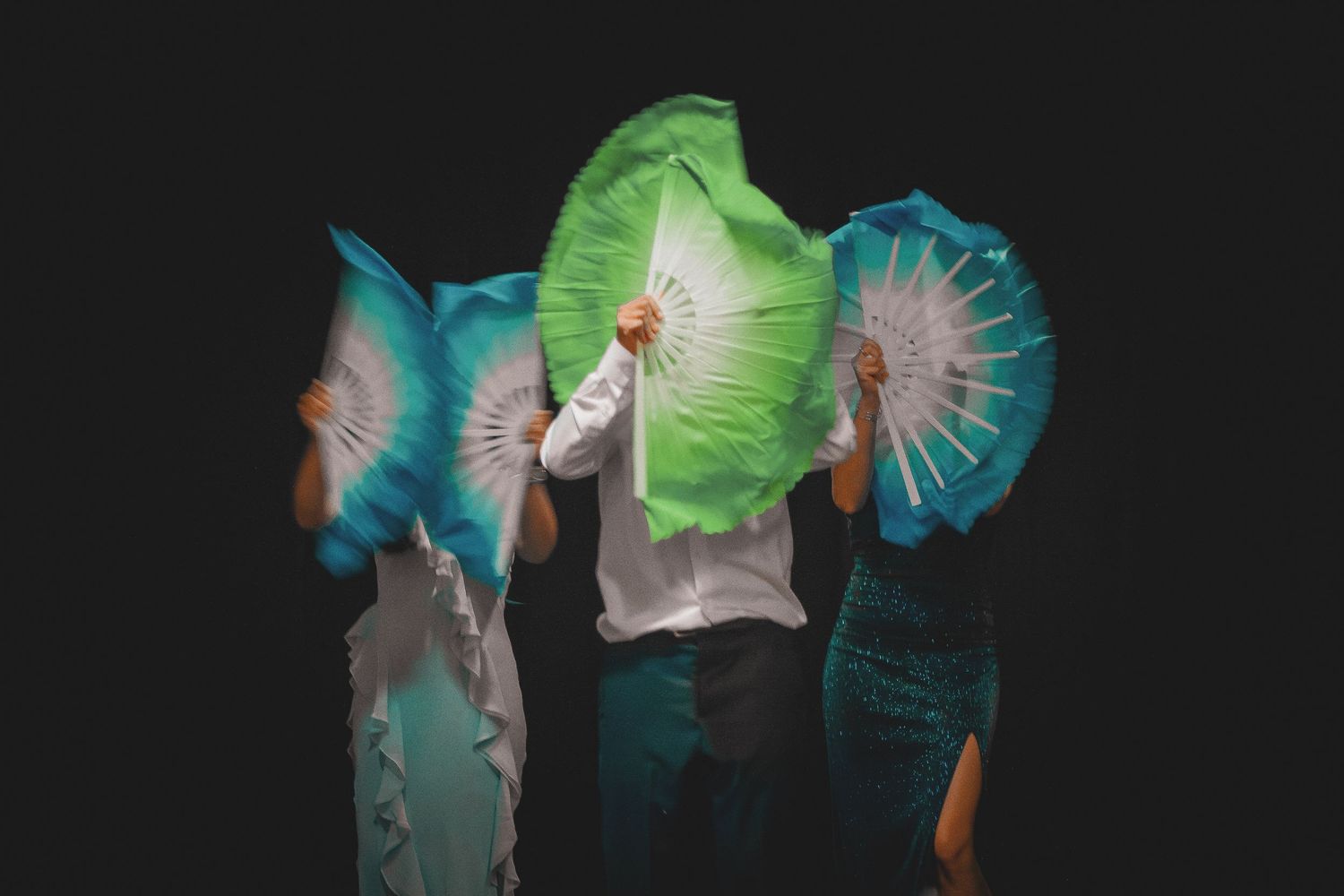 Three performers hold large colorful silk fans in green and blue, obscuring their faces on stage.