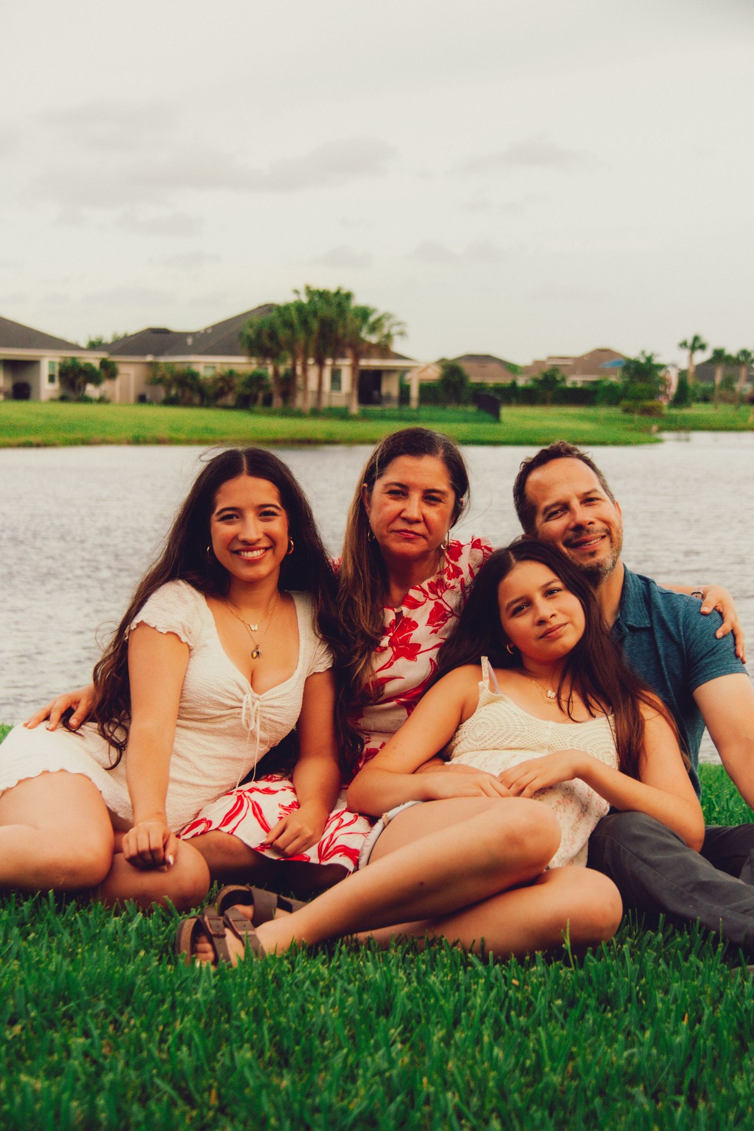 A family of four sits together smiling on green grass beside a lake with homes in the background.