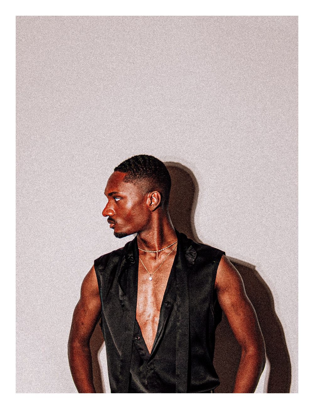 Side profile of a young Black man in an open sleeveless black satin vest with chain necklace in studio.