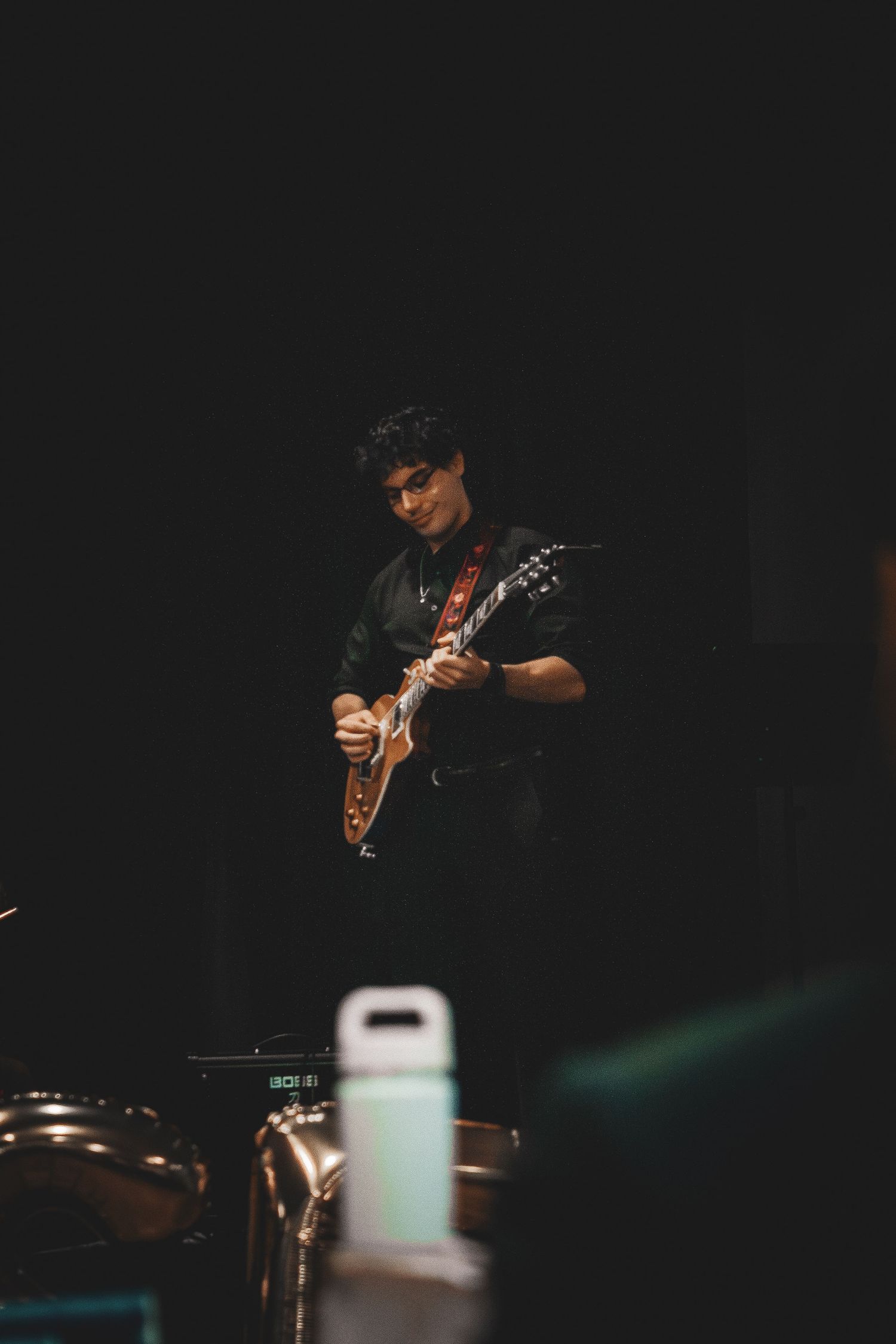A guitarist performs on a dark stage wearing all black while playing an electric guitar.