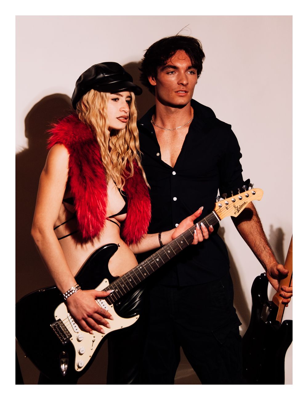 A blonde woman in a red fur vest and leather cap poses with a dark-haired man holding electric guitars.