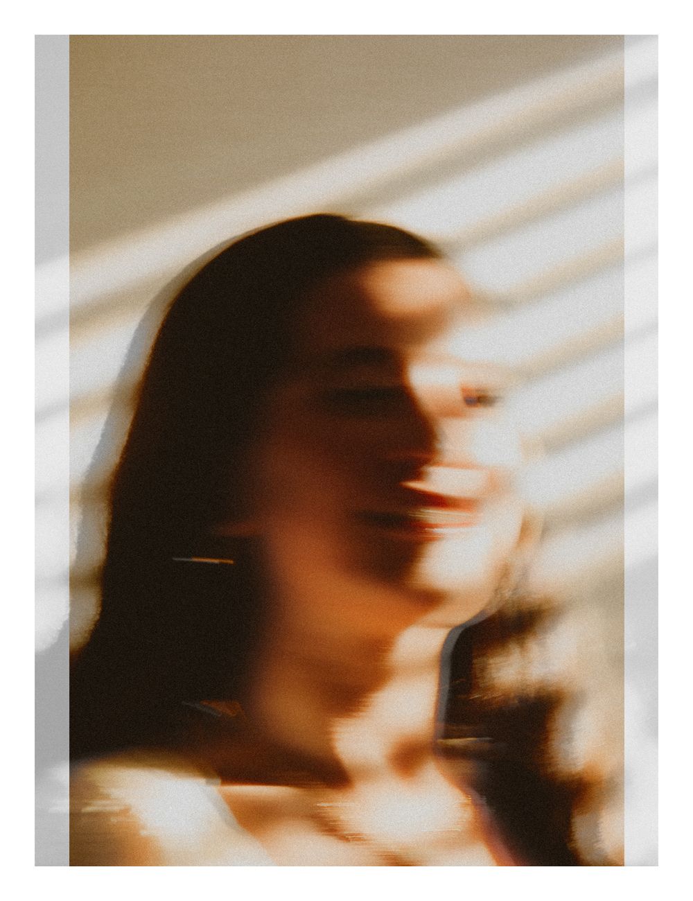 A blurred artistic portrait of a smiling woman bathed in warm sunlight through window blinds.