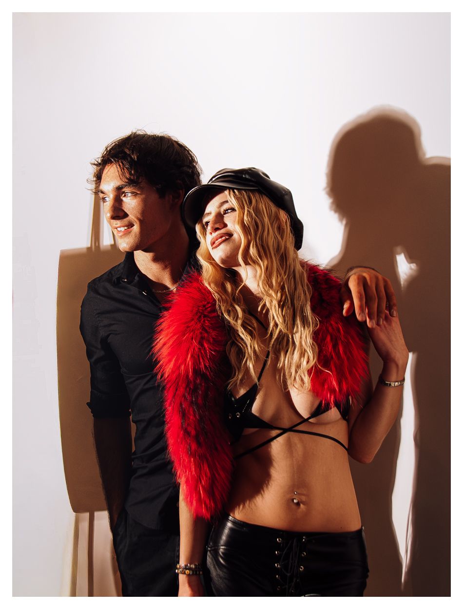 A smiling man in black shirt and woman in red fur stole and leather cap pose together with shadows behind them.