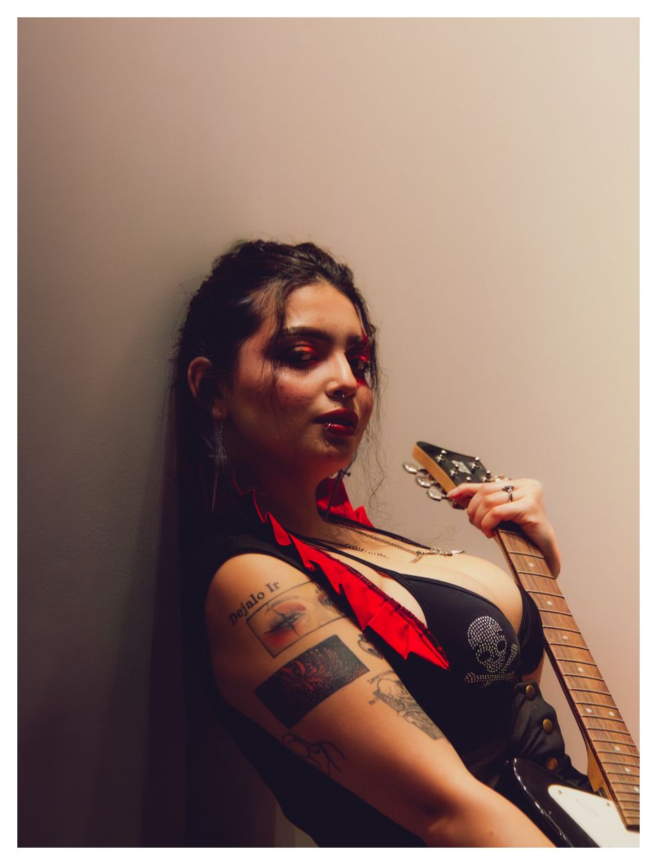 Tattooed woman in black top holding a guitar neck, wearing a red bow tie, leaning against a wall.