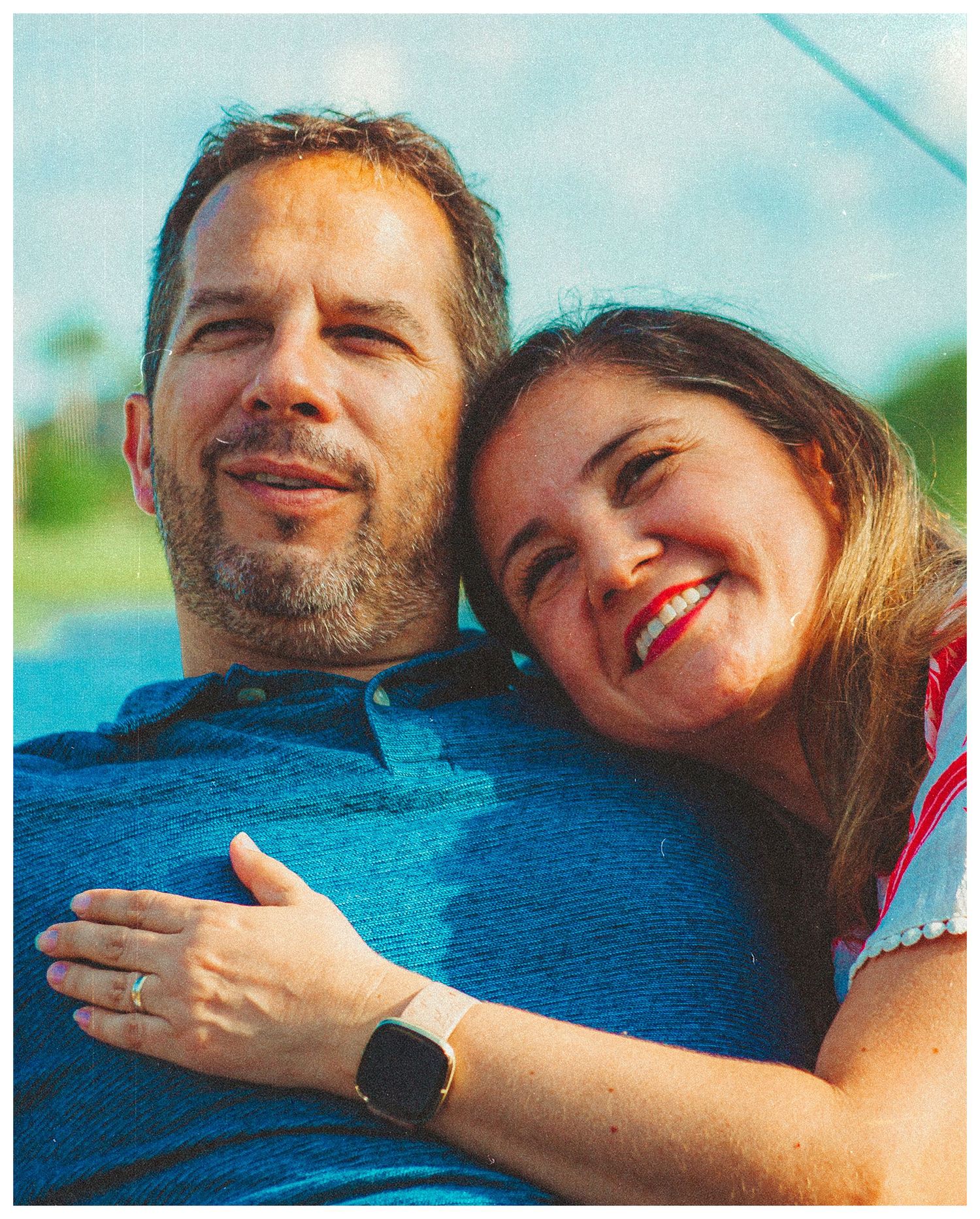 Smiling couple embracing outdoors, man in blue shirt and woman in red top near a body of water.