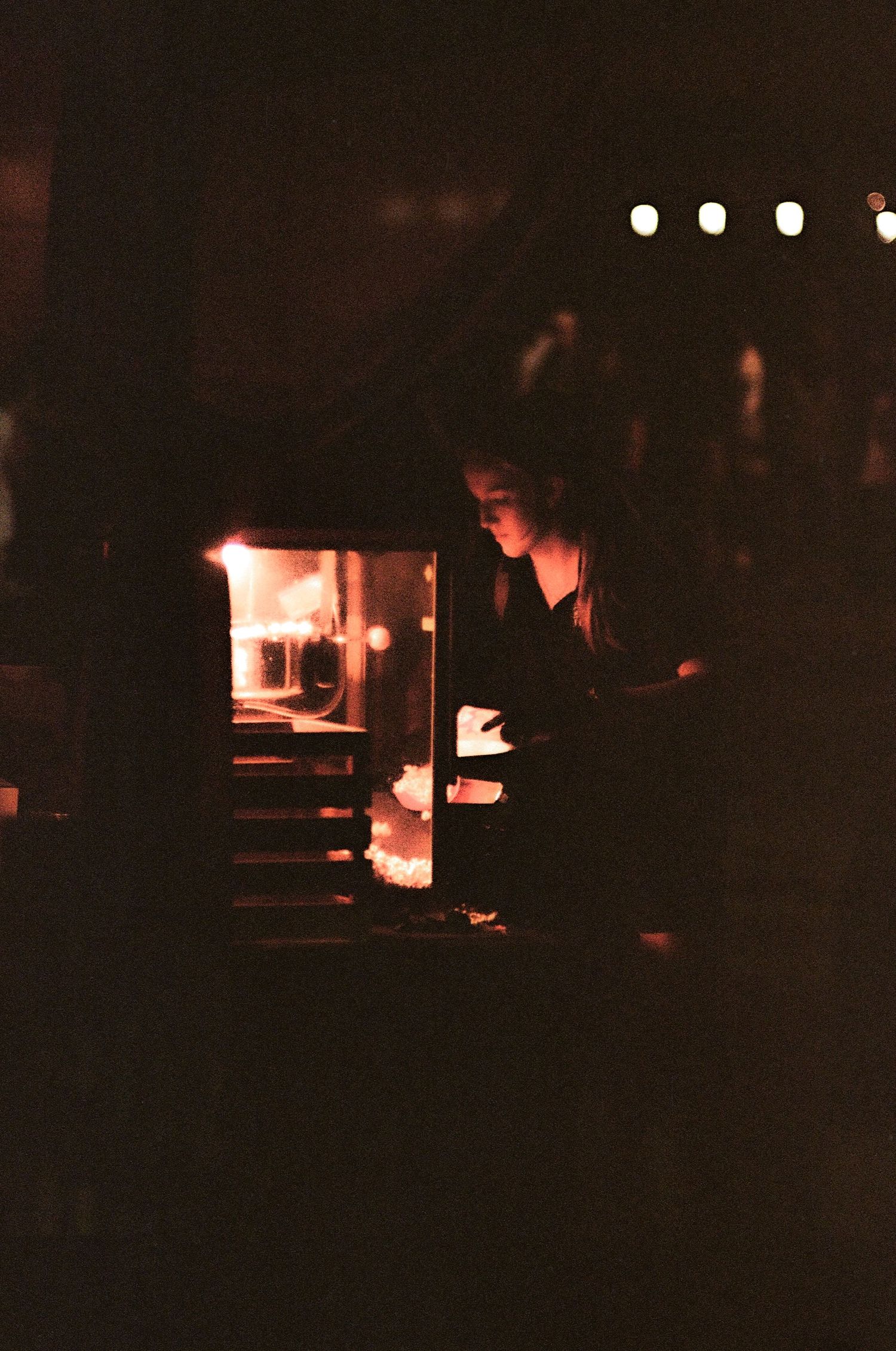 A woman works by glowing firelight in a dark, moody film photograph with bokeh lights behind.