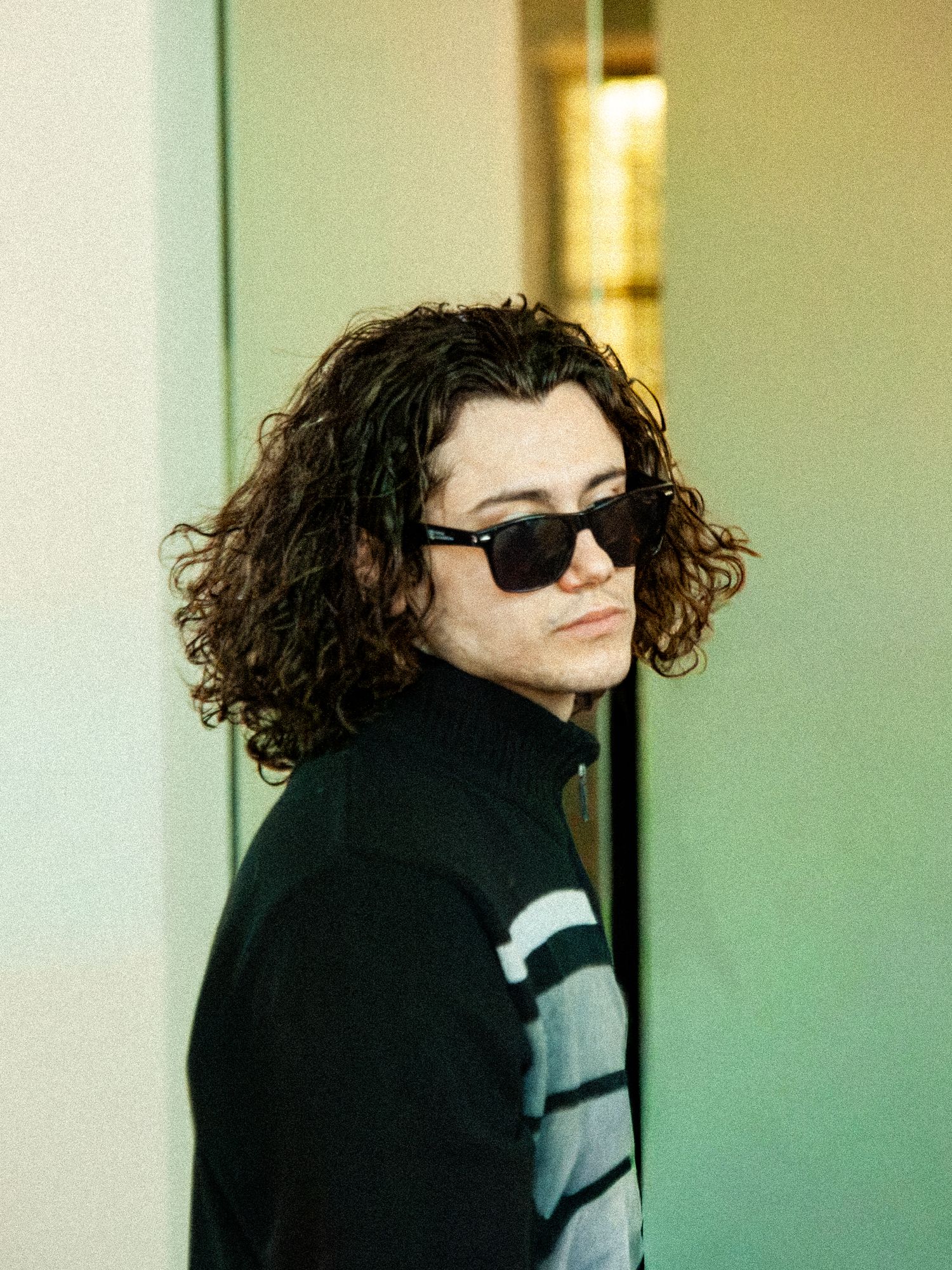 Young man with curly hair wearing sunglasses and black turtleneck sweater outdoors.