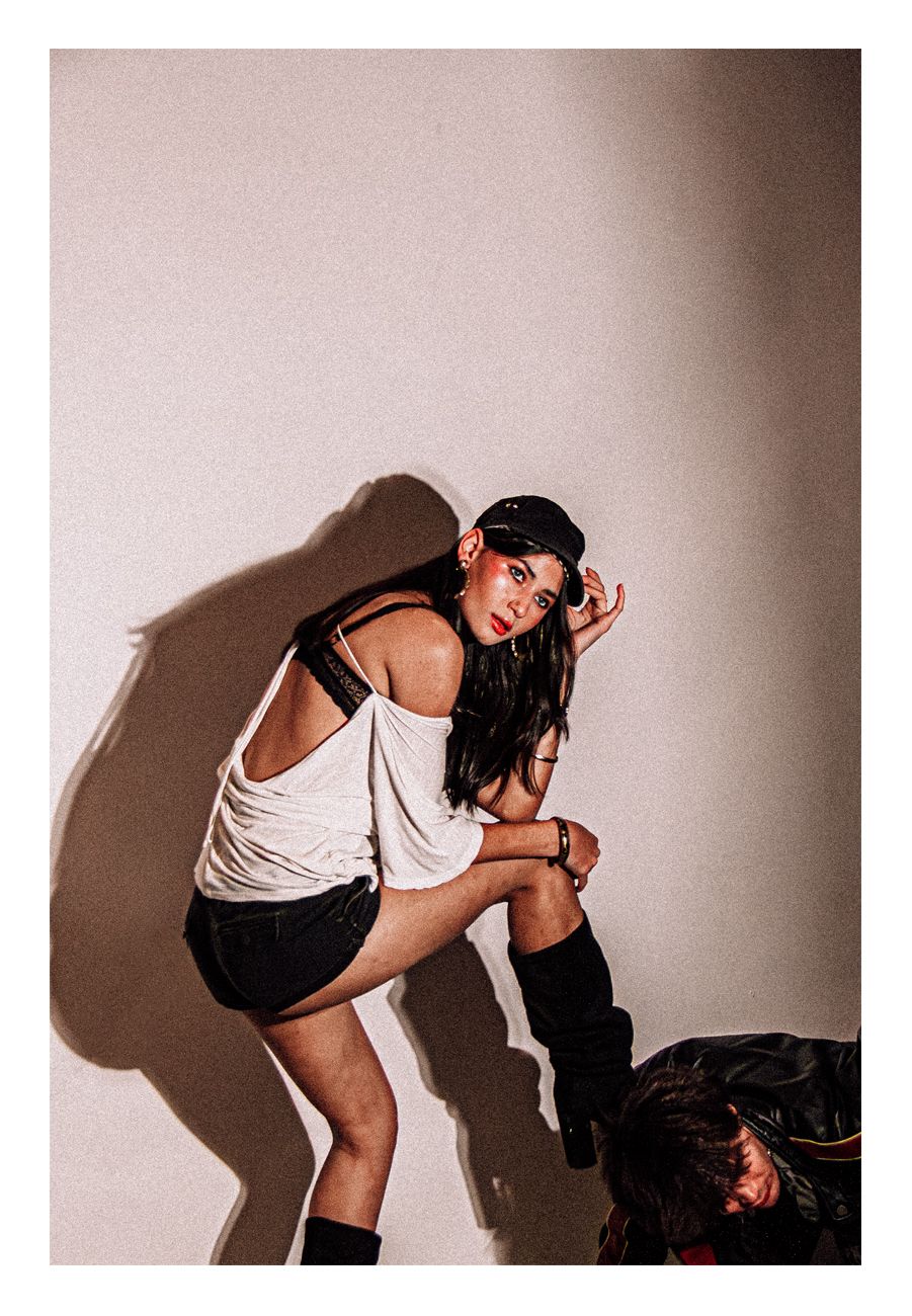 Woman in white tank and black shorts leans forward playfully in a studio setting.