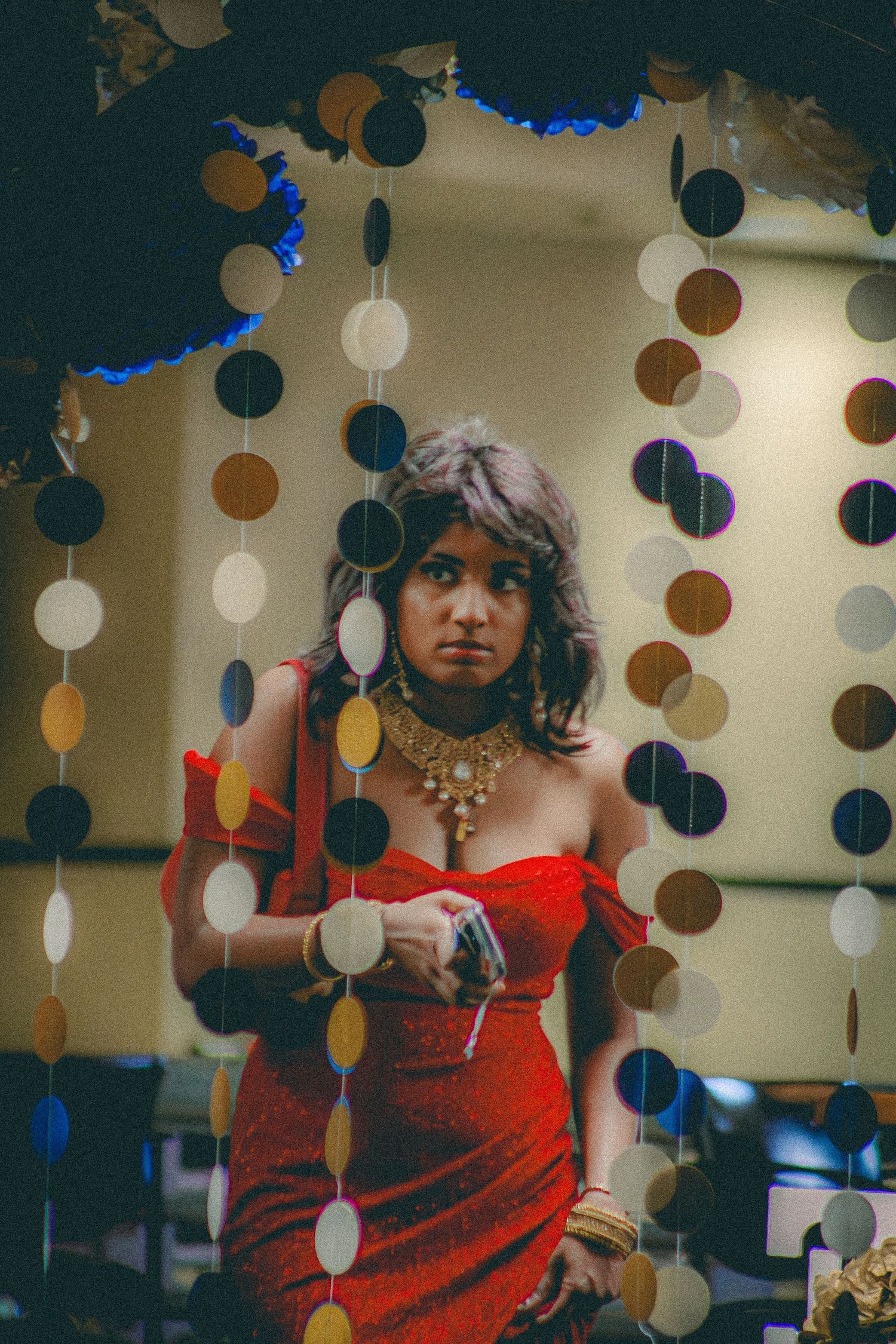 A woman in a red off-shoulder dress adorned with gold jewelry poses near circular garland decor.
