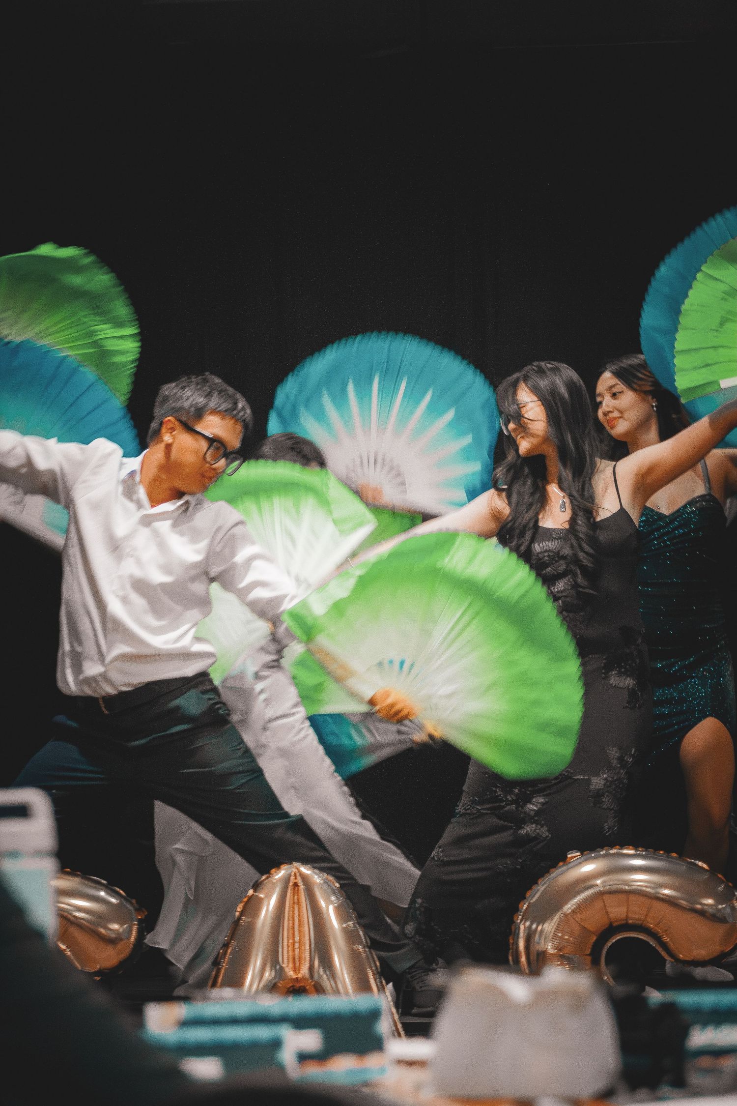 Group of young Asian dancers performing with colorful green and blue silk fans on a dark stage.