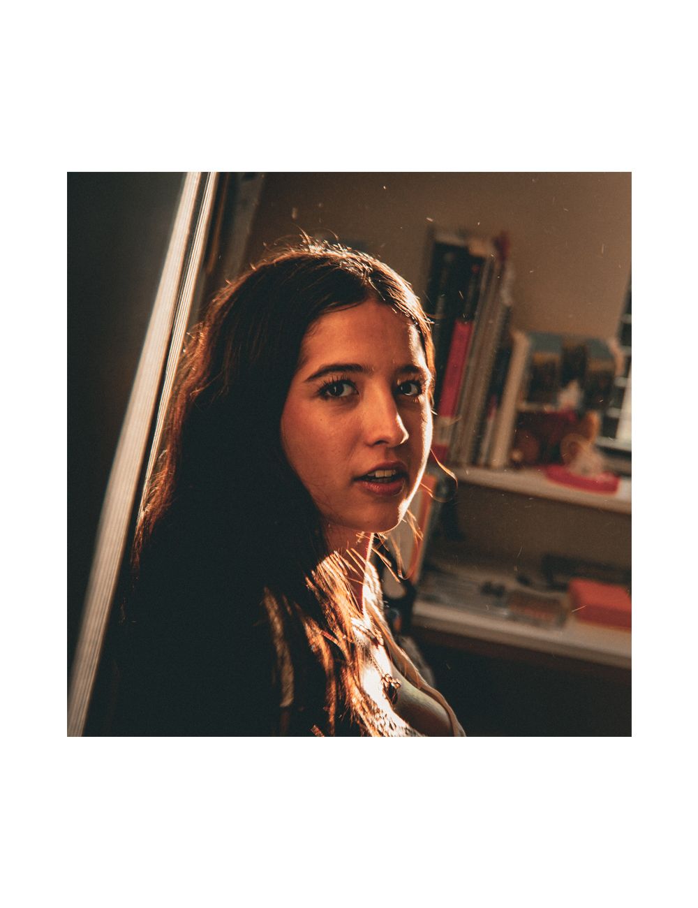 Young woman with long dark hair lit by warm golden light standing near a window in a cozy room.