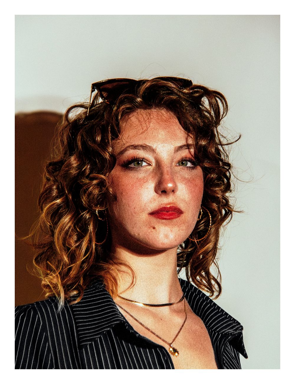 Curly-haired woman with red lips, freckles, and sunglasses in a striped blazer.