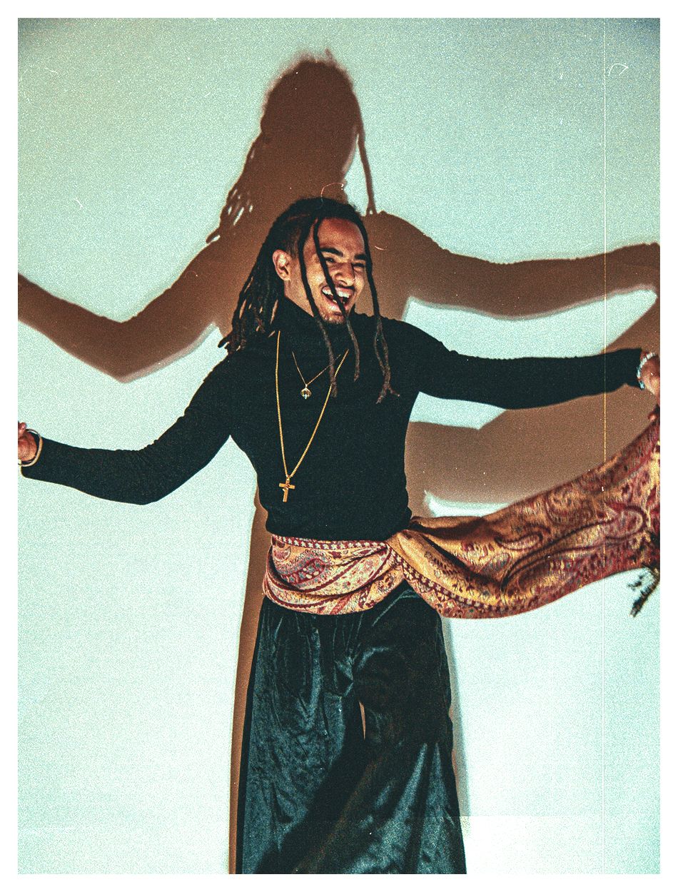 A person with dreadlocks dances joyfully in a black top and ornate sash skirt.