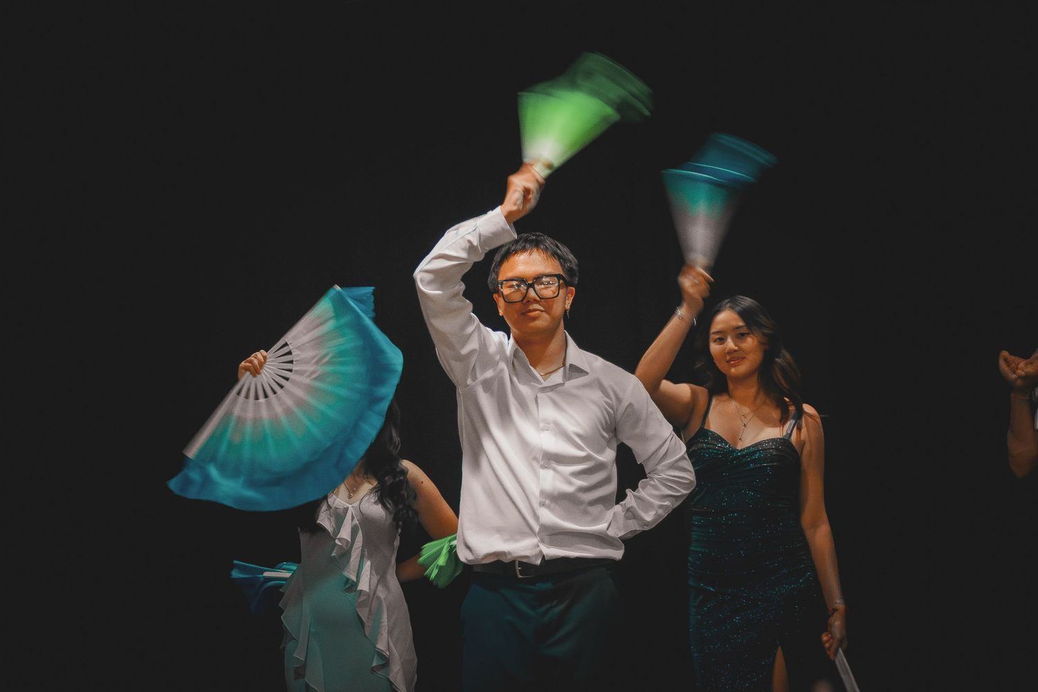 Young Asian man in white shirt dancing energetically with green and teal silk fans on dark stage.