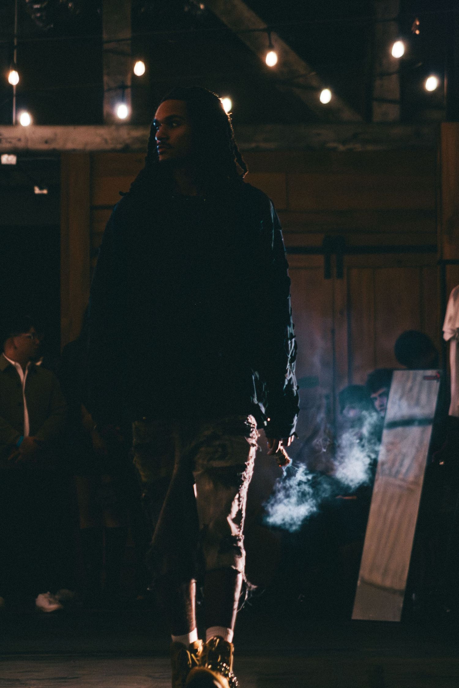 A man holding a smoking torch stands in a dimly lit barn with string lights overhead.
