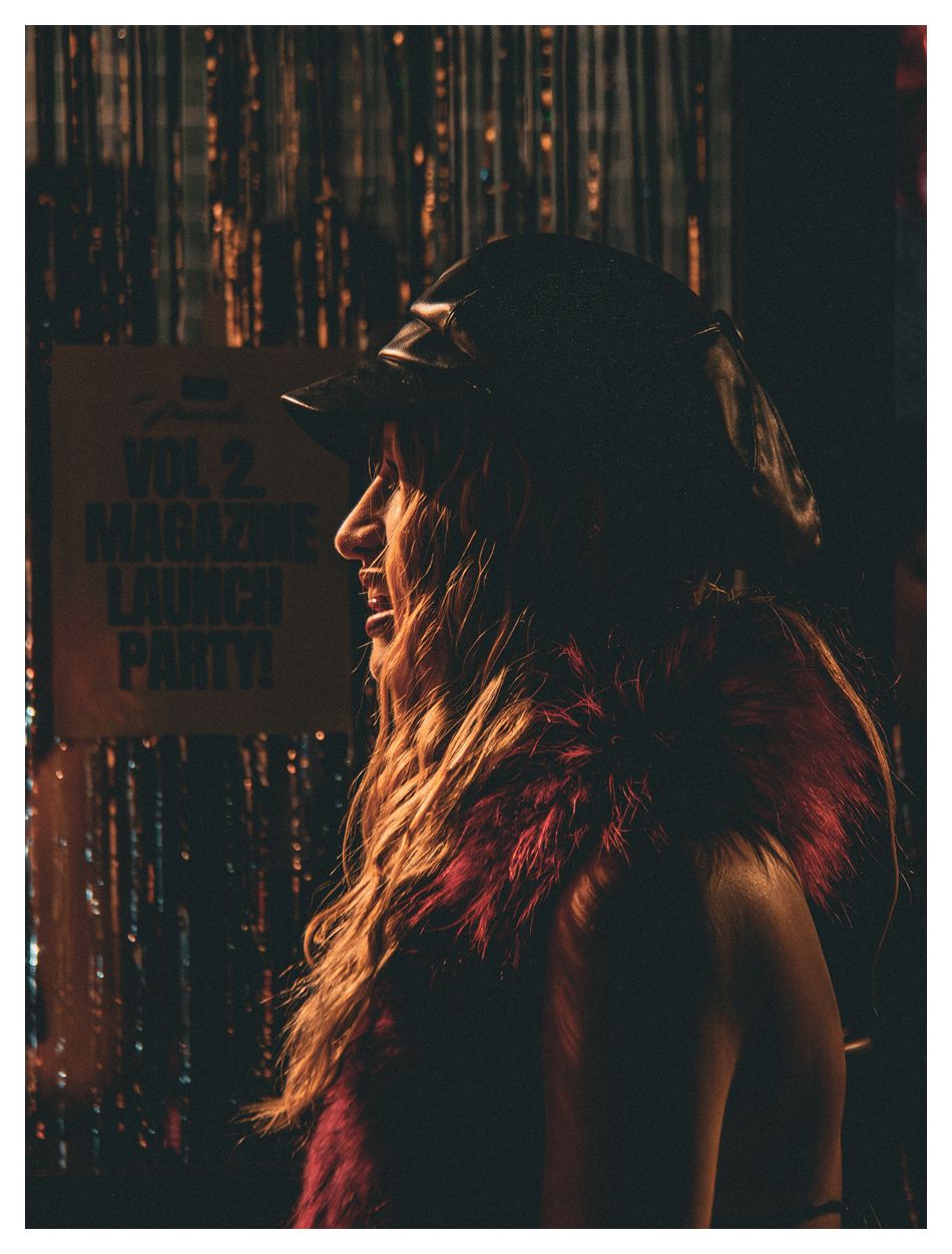 Side profile of a smiling woman with long hair wearing a cap and red fur at a Vol. 2 Magazine Launch Party.