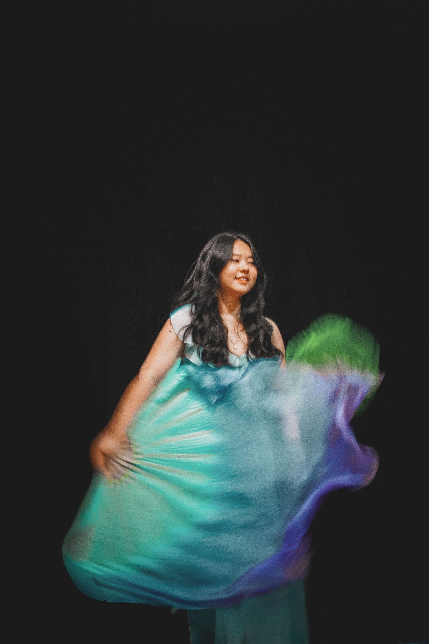 Young Asian woman in turquoise dress smiling while performing flowing silk fan dance on dark stage.