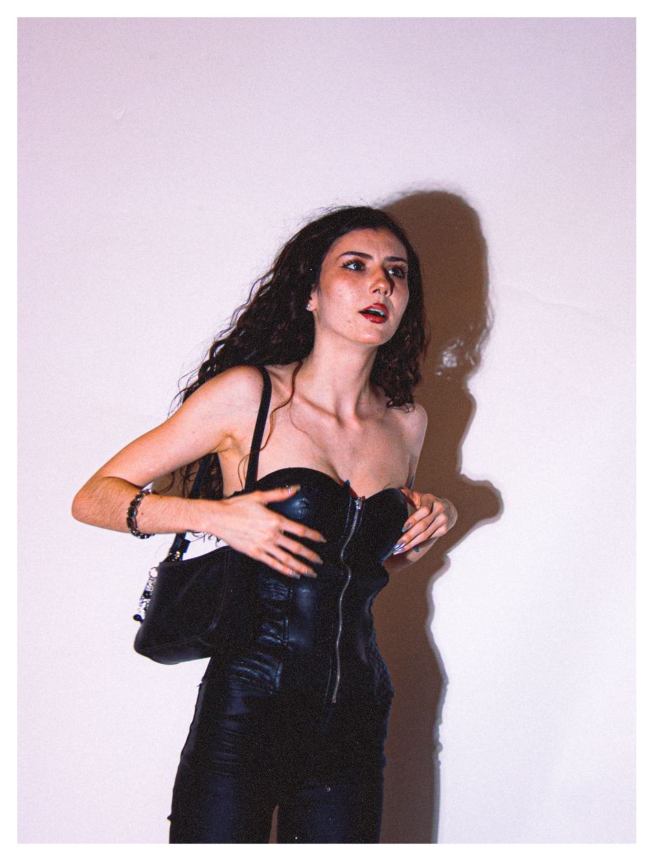 Woman in black leather bustier and skirt poses dramatically against white wall.